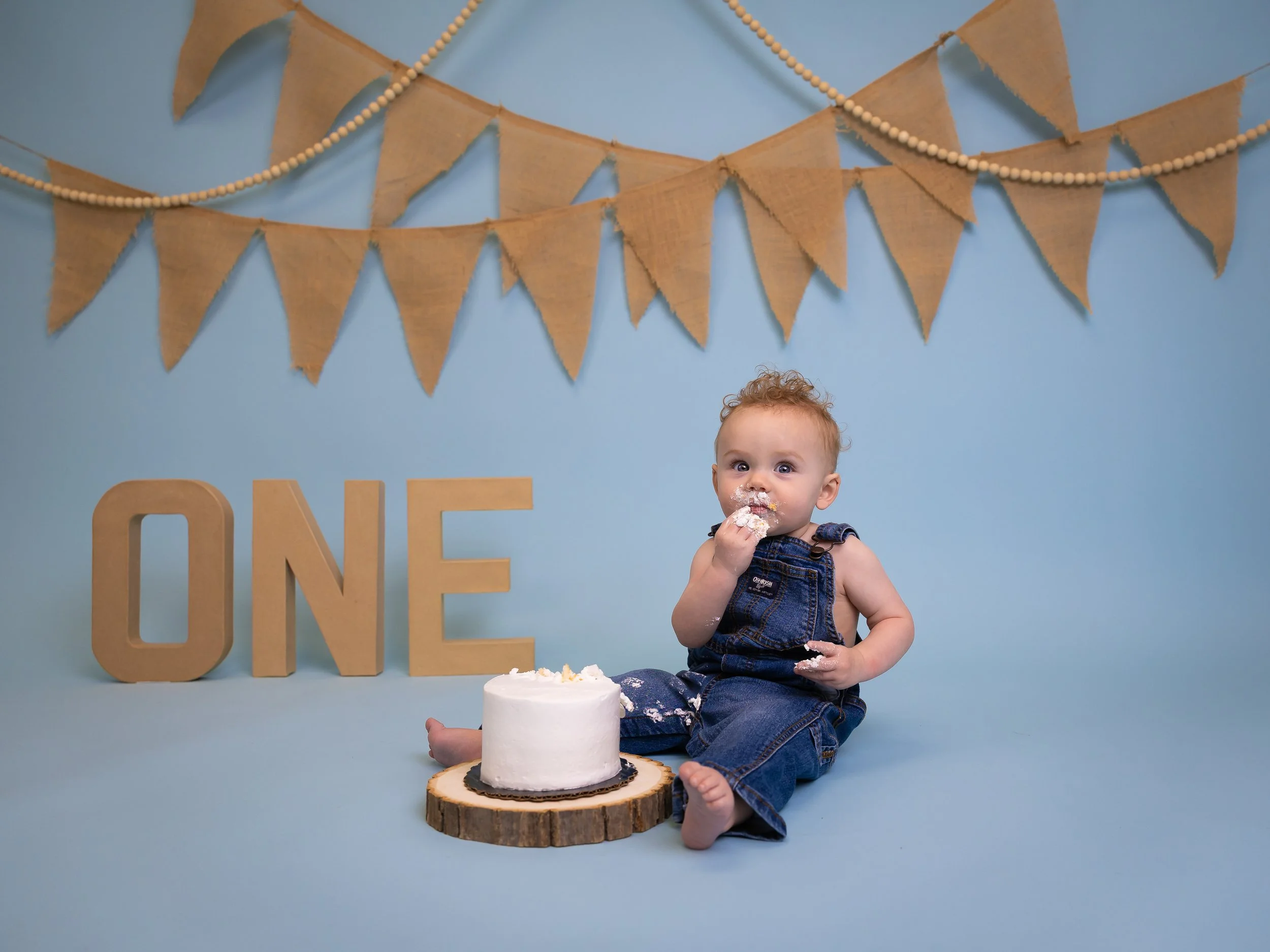 A young child celebrating their first birthday, sitting on the floor in denim overalls. In front of the child is a small white cake on a wooden slab. A light blue backdrop with tan-colored bunting and the word "ONE" in tan letters marks the birthday