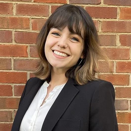 A young woman with shoulder-length brown hair, smiling, wearing a black blazer and a white blouse, standing in front of a brick wall.