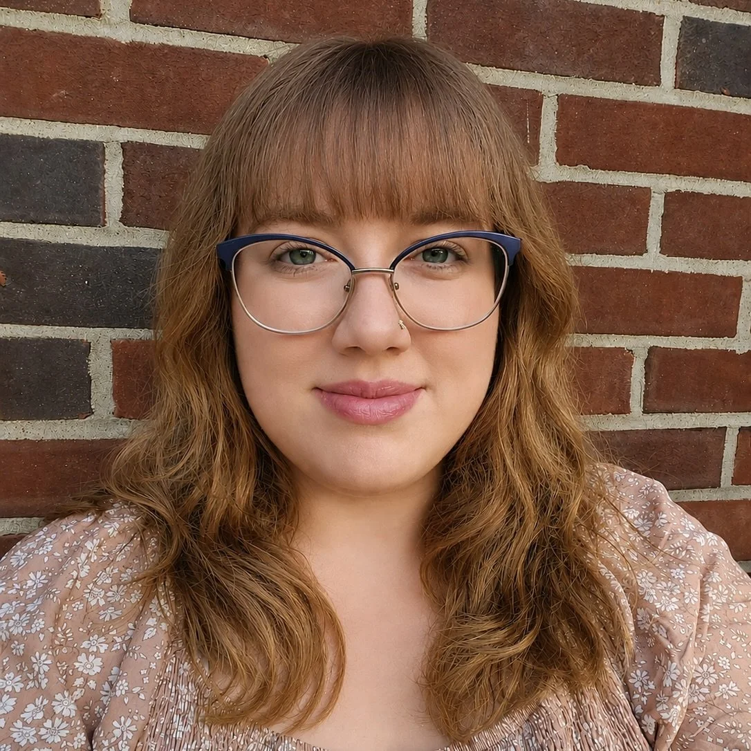 A woman with red hair and glasses taking a selfie in front of a brick wall.