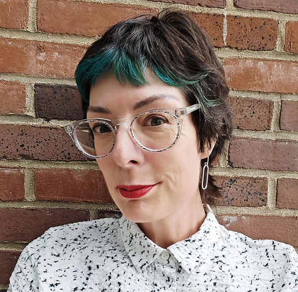 Shannon, a woman with short hair, with teal highlights, wearing clear glasses, red lipstick, a white patterned shirt, and silver earrings, standing against a brick wall.