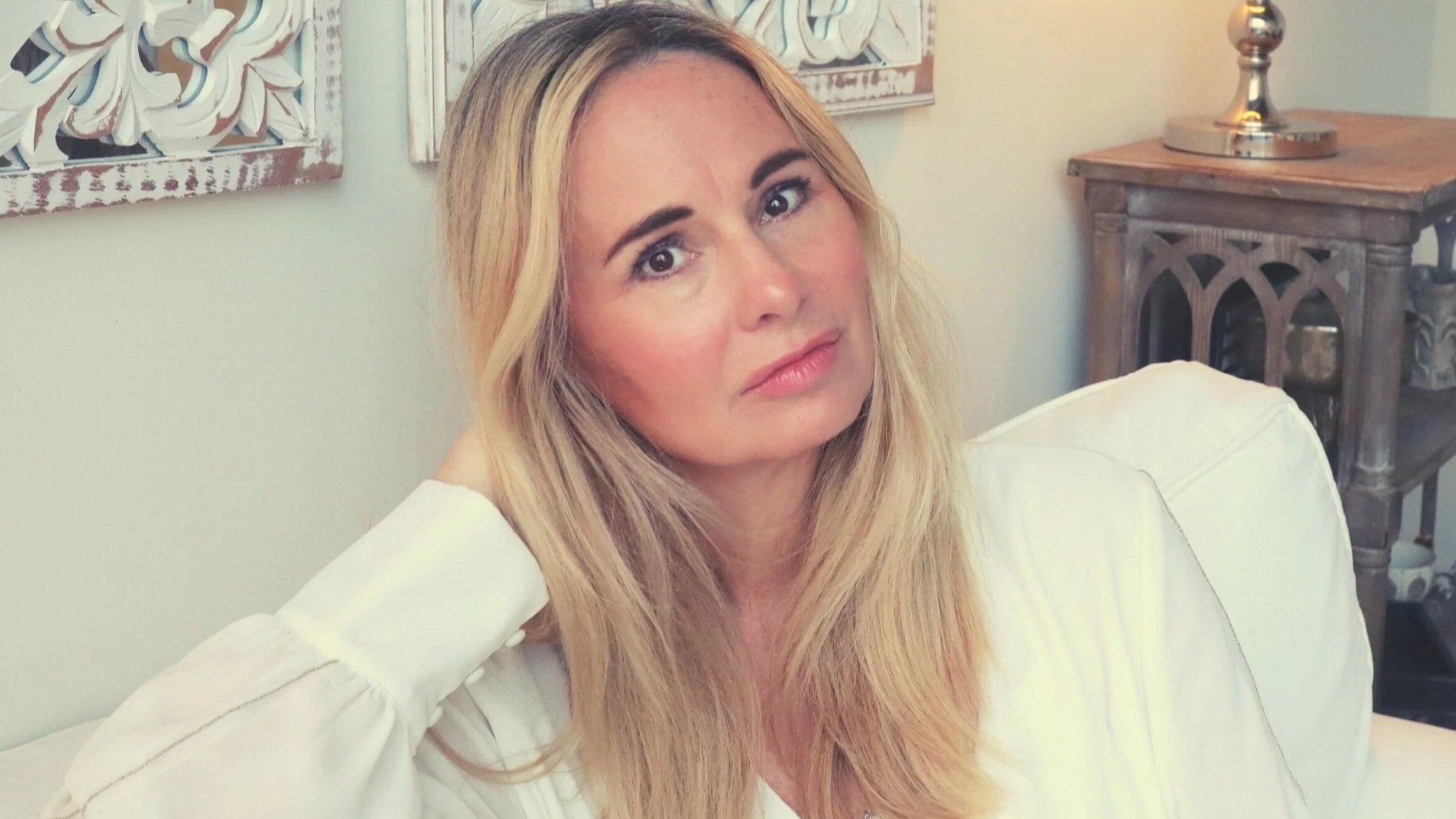 A woman with long blonde hair wearing a white top sitting indoors with decorative art on the wall behind her and a wooden table with a lamp to the right.