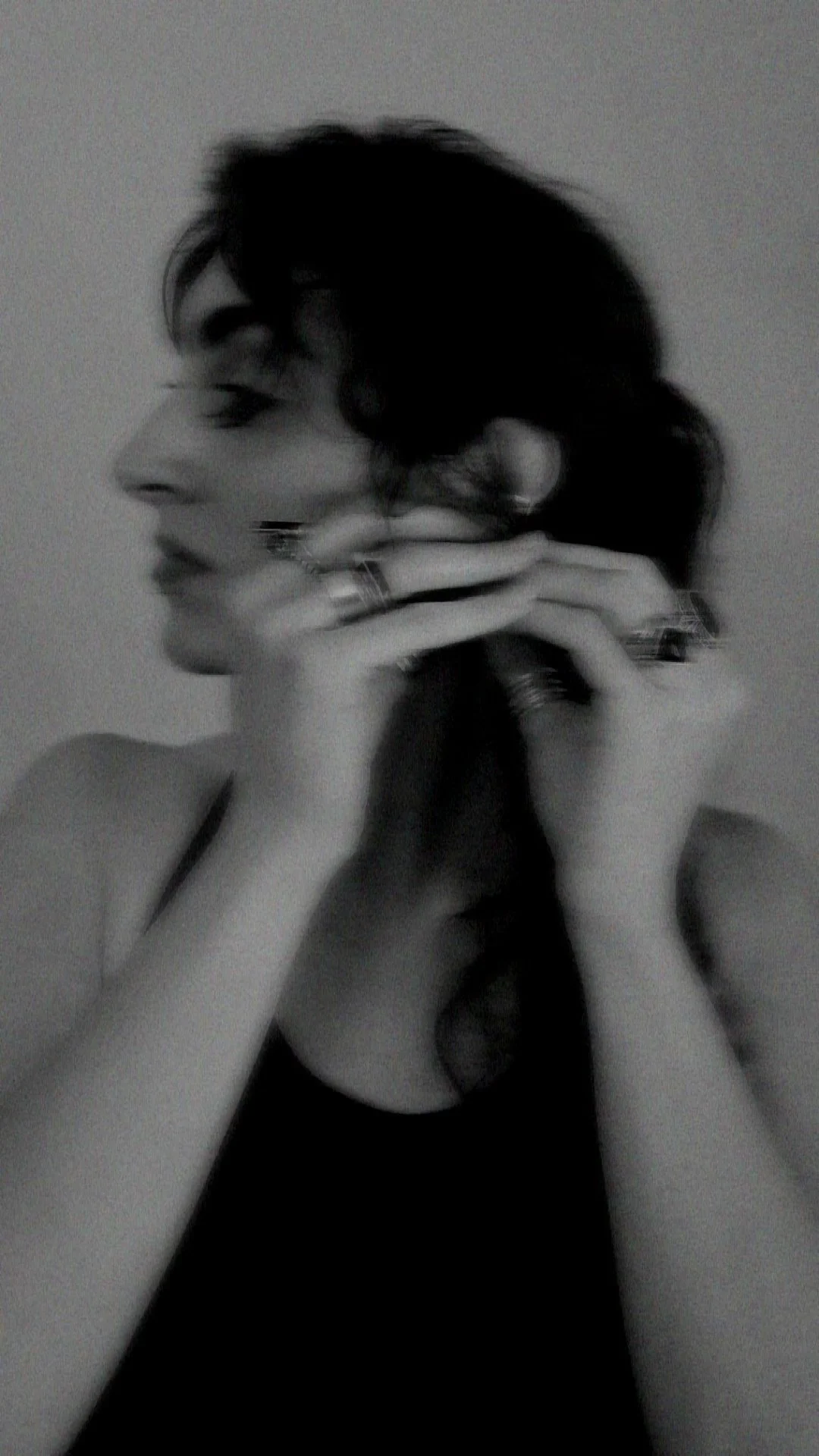 Black and white selfie of a woman with short dark hair, wearing rings and a sleeveless top, with her eyes closed and hands resting on her face.