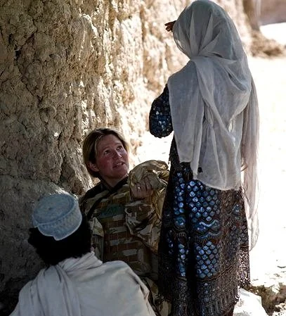 Rosie Stone, engaged in a conversation outdoors in Afghanistan, near a rocky wall; the woman is standing with her back to the camera, wearing a headscarf and patterned dress, while the man facing her has a beard and is wearing a patterned shirt.