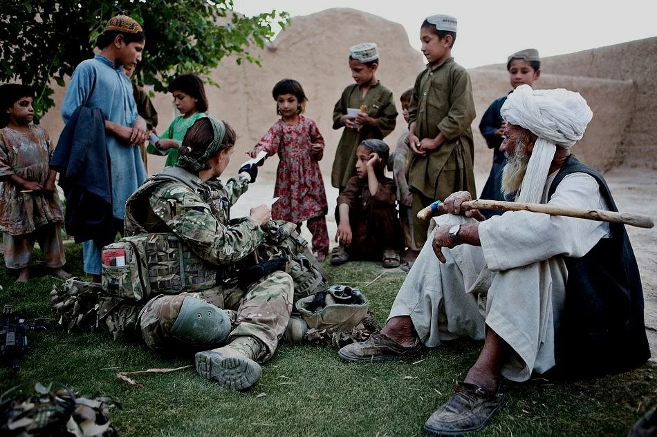 A military soldier and an elderly man in Afghanistan in traditional clothing and turban sitting outdoors surrounded by children in a rural setting.