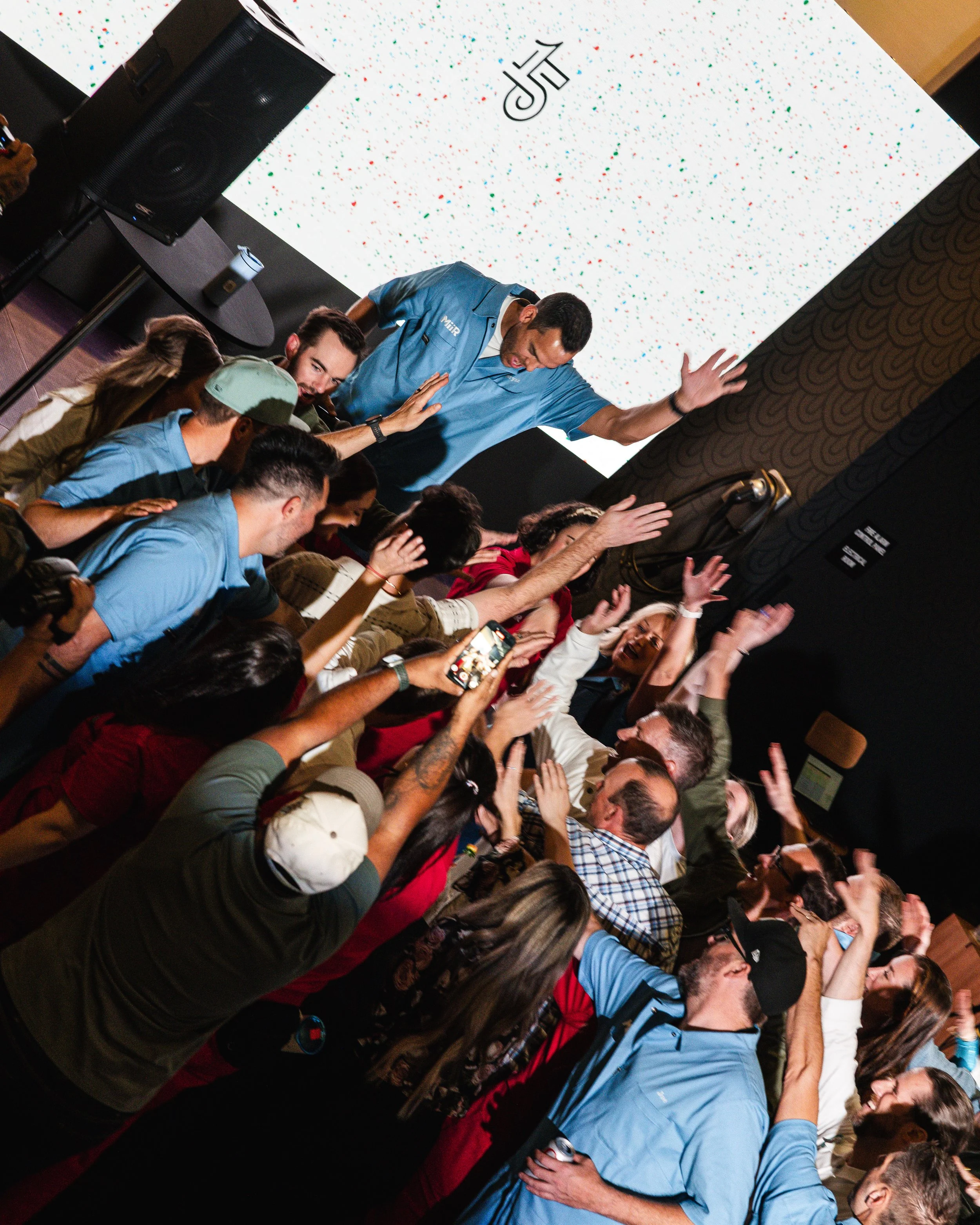 Celebration or party scene with a group of people reaching out towards a man on stage, some taking photos, in a room with a large screen displaying colorful speckles and initials 'JJK'.