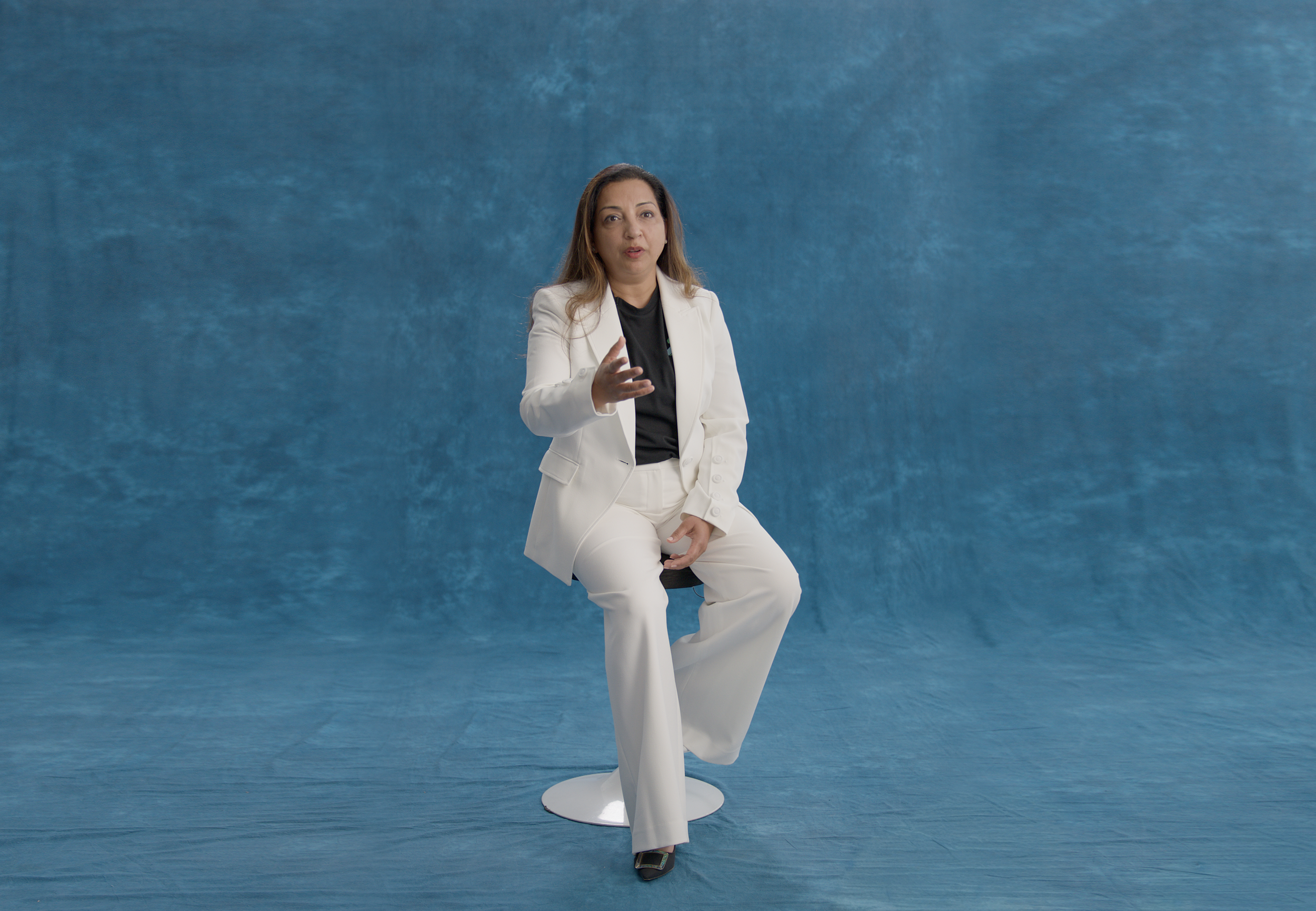 Woman in a white suit sitting on a round stool with a blue background.