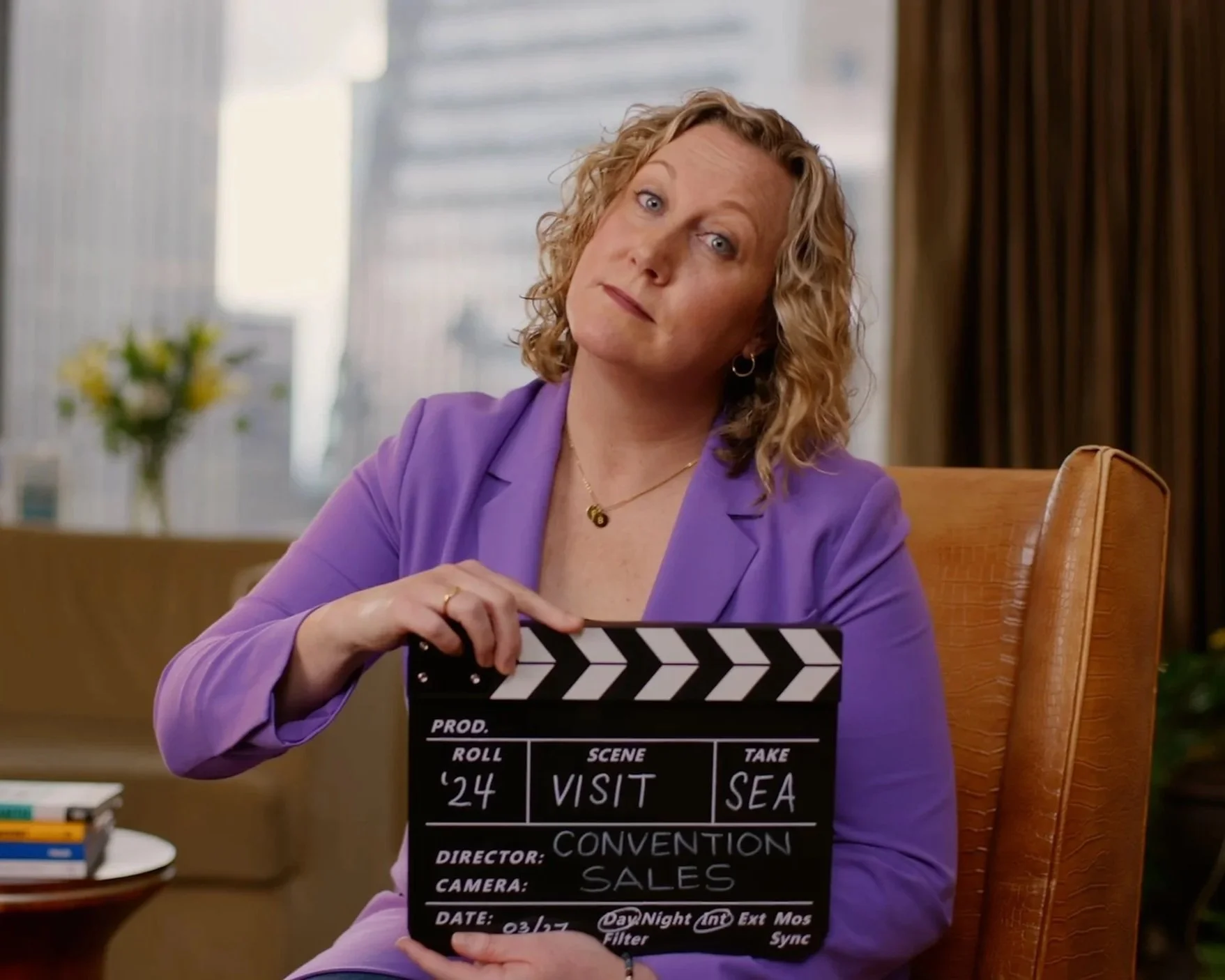 A woman with curly blonde hair wearing a purple blazer and gold jewelry holding a film slate that reads 'Visit SEA' in an office setting with a blurred cityscape background.