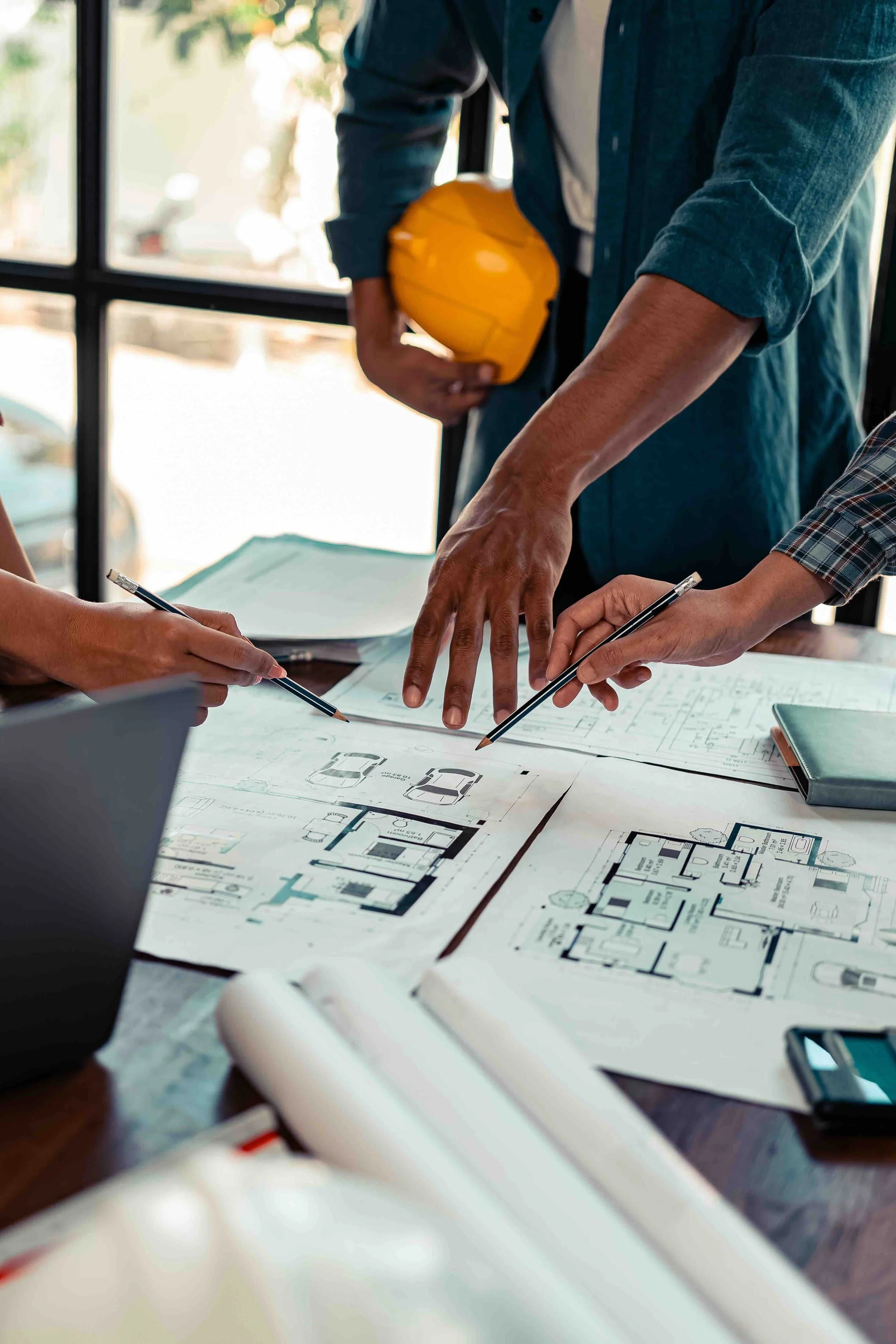 People working on architectural or engineering blueprints spread out on a table, with one person pointing at the plans and others holding pens, in a bright room with large windows.