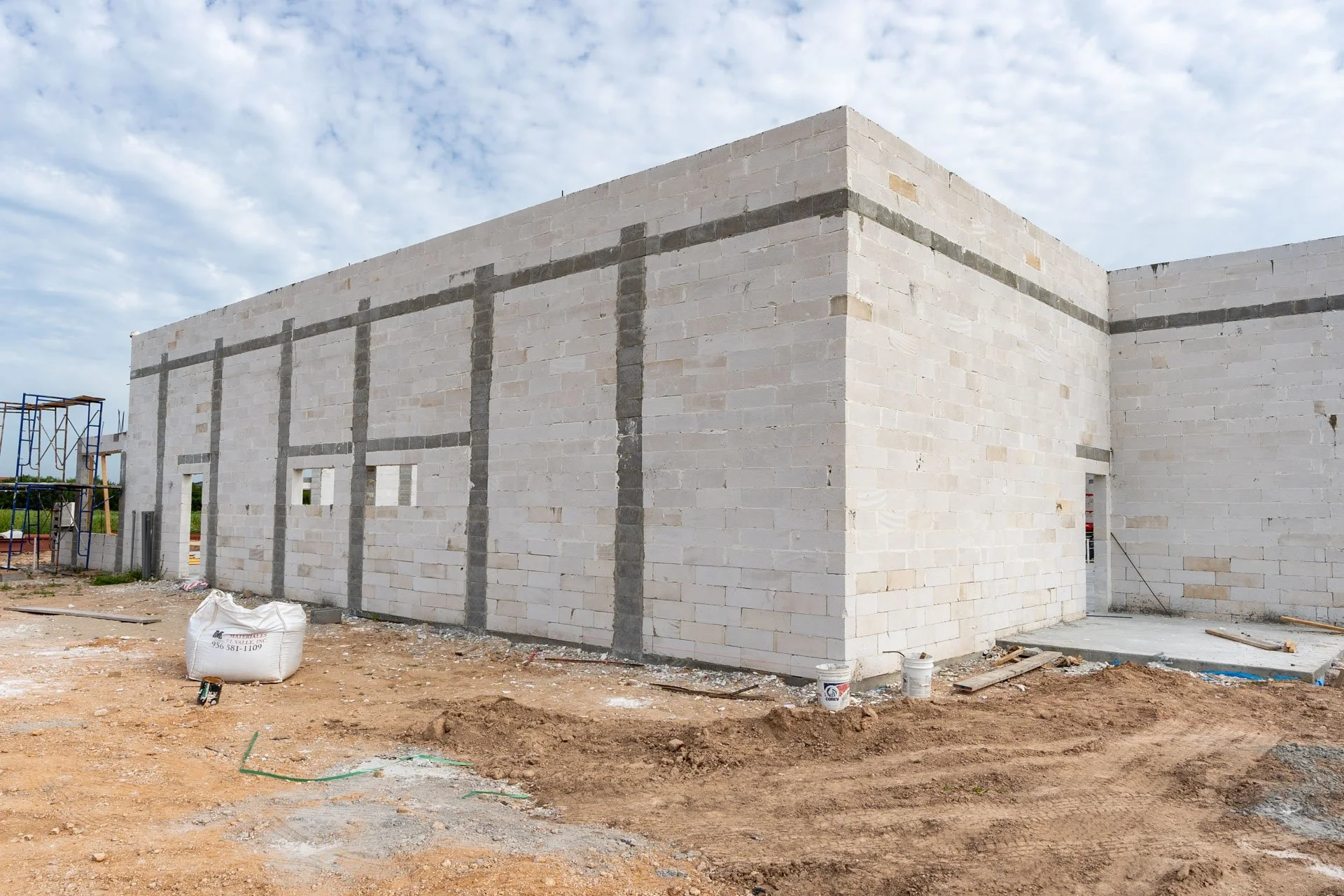 A building under construction with white cinder block walls and visible gray concrete mortar lines, with a partly cloudy sky overhead.