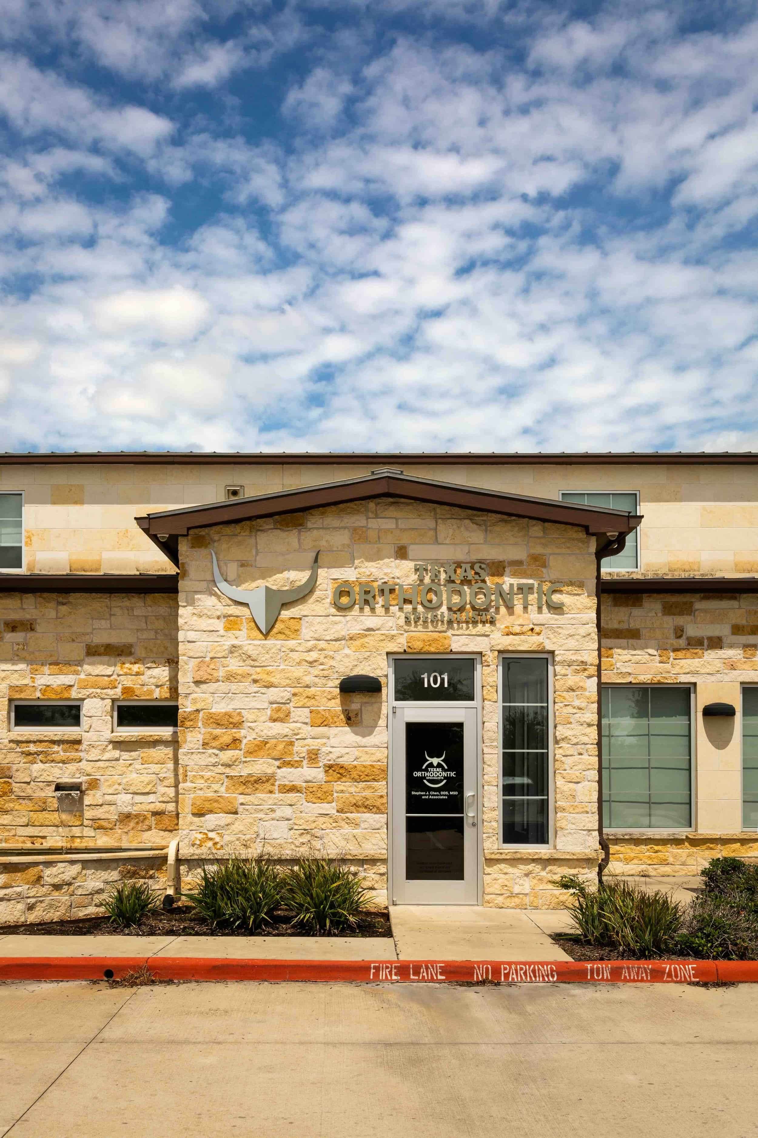 Front of a stone building with a sign that reads 'Texas Orthodontic Specialists'. The building has a glass door with the same logo and text, and the street has a red curb with fire lane markings.