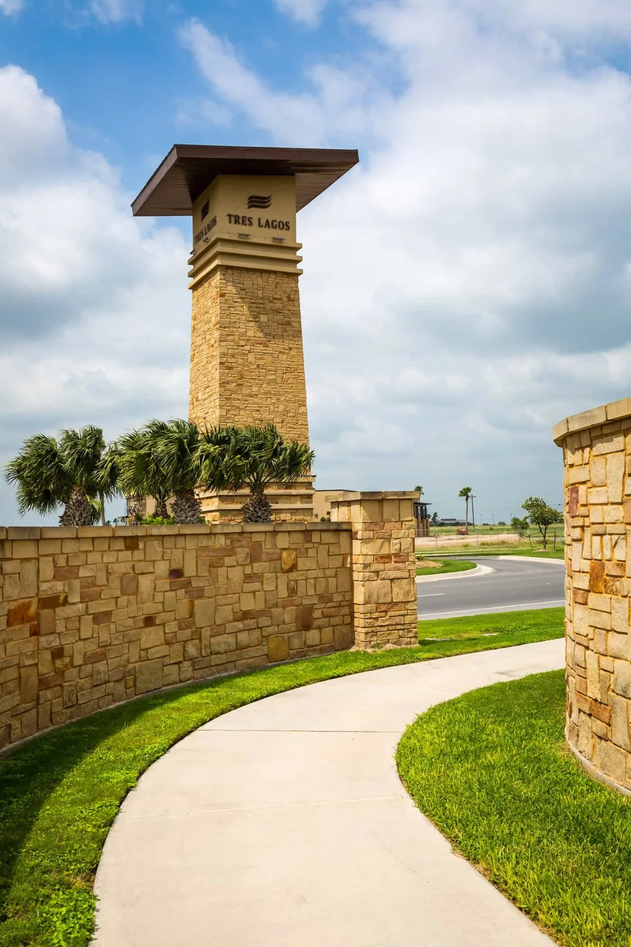 Stone wall and curved sidewalk leading to a large sign with palm trees in front. The sign reads 'Tres Lagos' and is part of a lamppost or tower structure against a cloudy sky in a suburban setting.