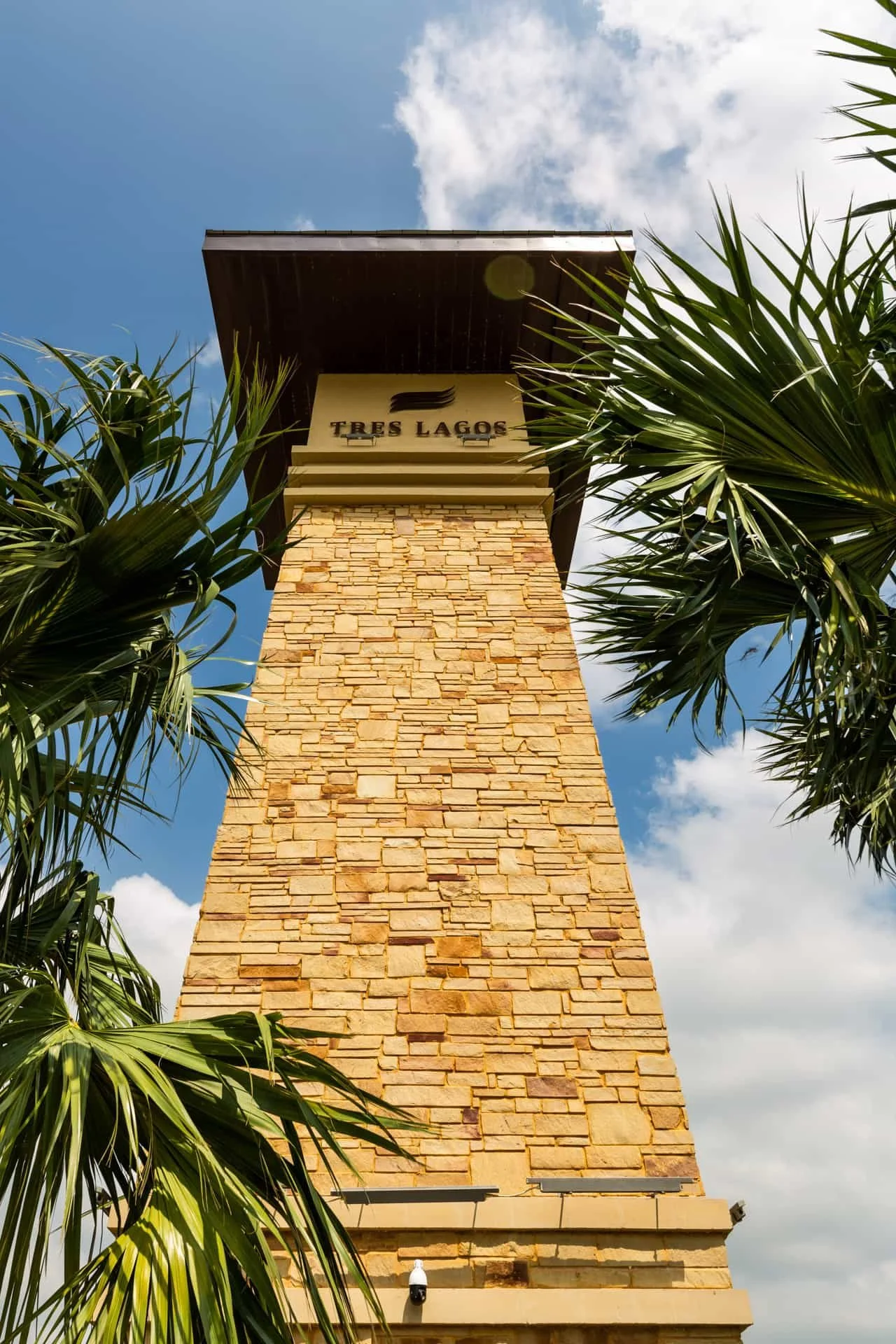 Tall stone tower with sign 'Tres Lagos' at the top, surrounded by palm trees and a partly cloudy sky.