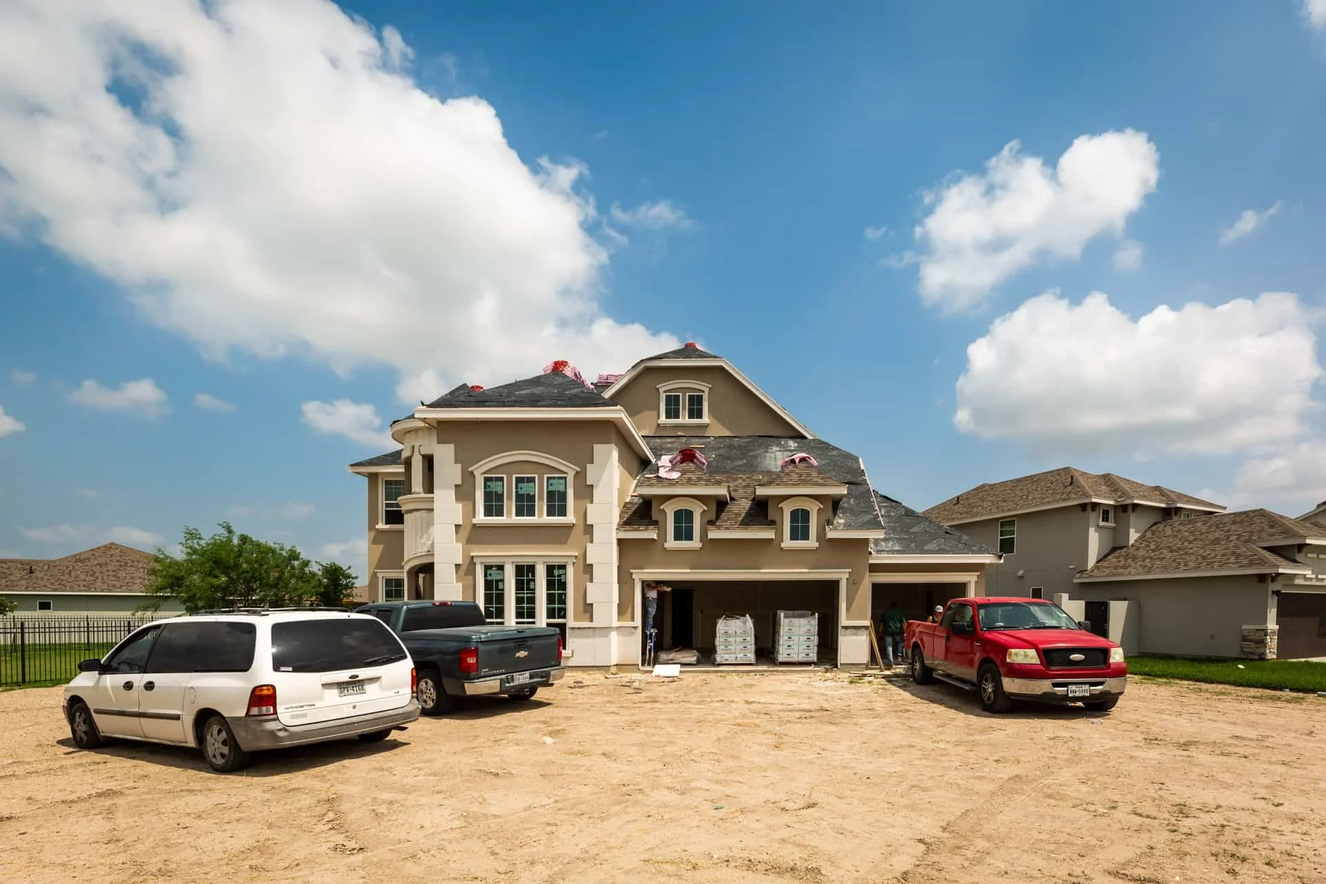 A large house under construction with a dirt driveway, several parked cars, and a blue sky with clouds.
