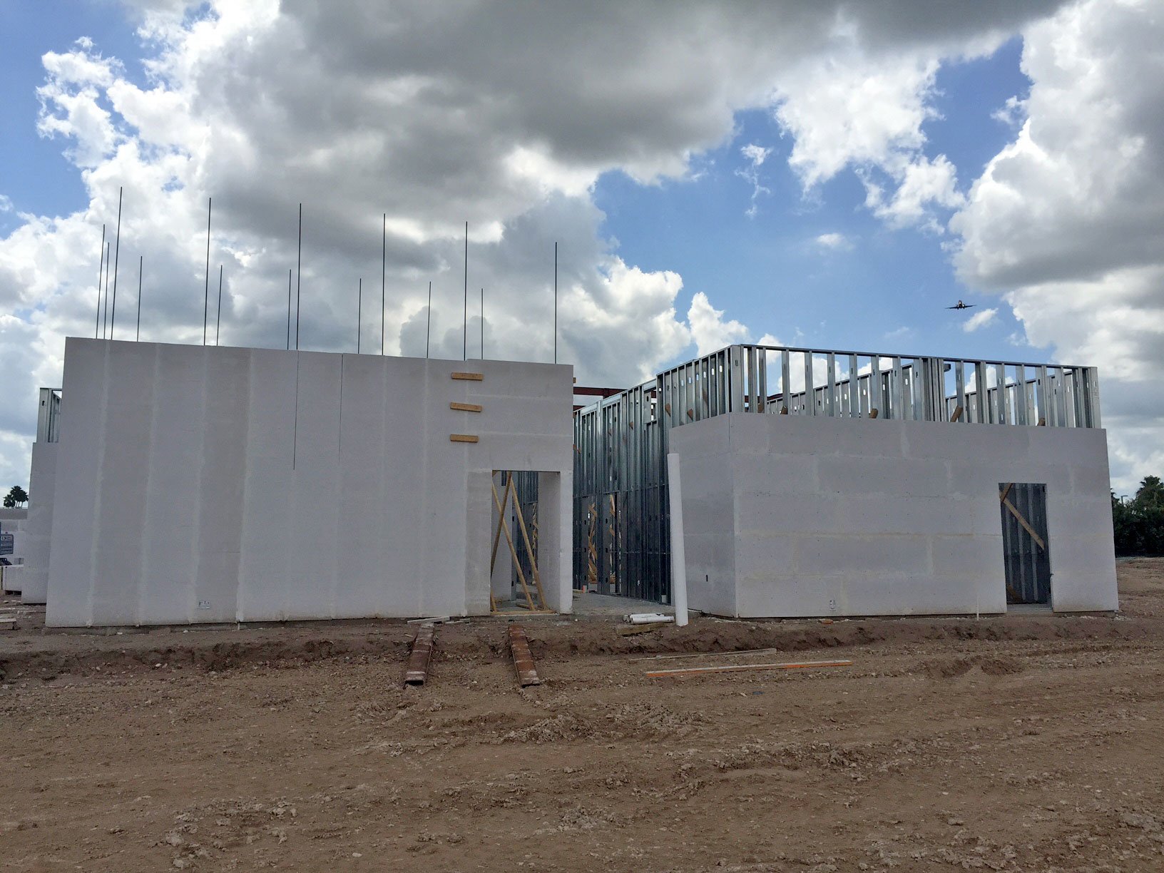 Under construction building with white walls and metal framing, situated on a dirt lots, under a partly cloudy sky, with an airplane flying in the background.