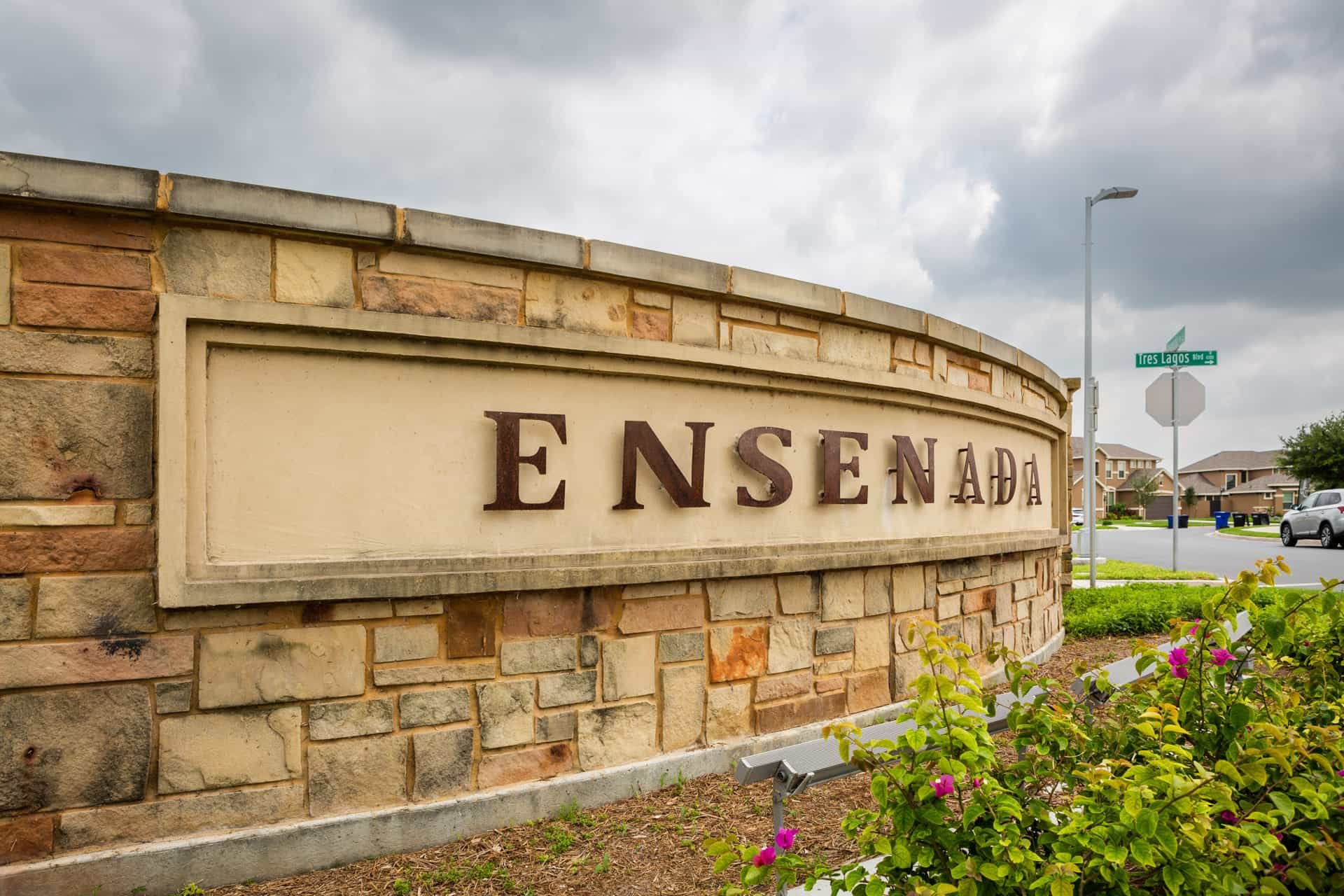 View of a brick neighborhood entrance sign that reads 'ENSEADA' with nearby street signs and houses in the background.