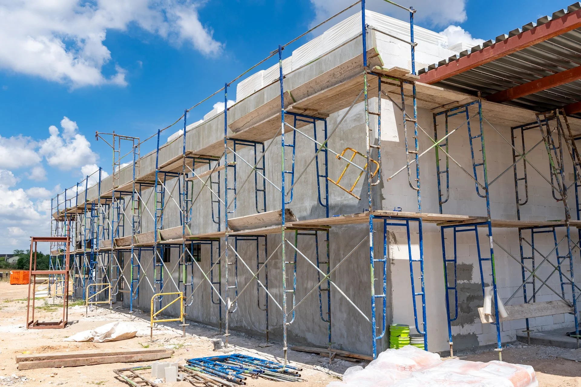 Construction site with scaffolding around a building under construction, with concrete walls, a metal roof, and blue sky with clouds in the background.
