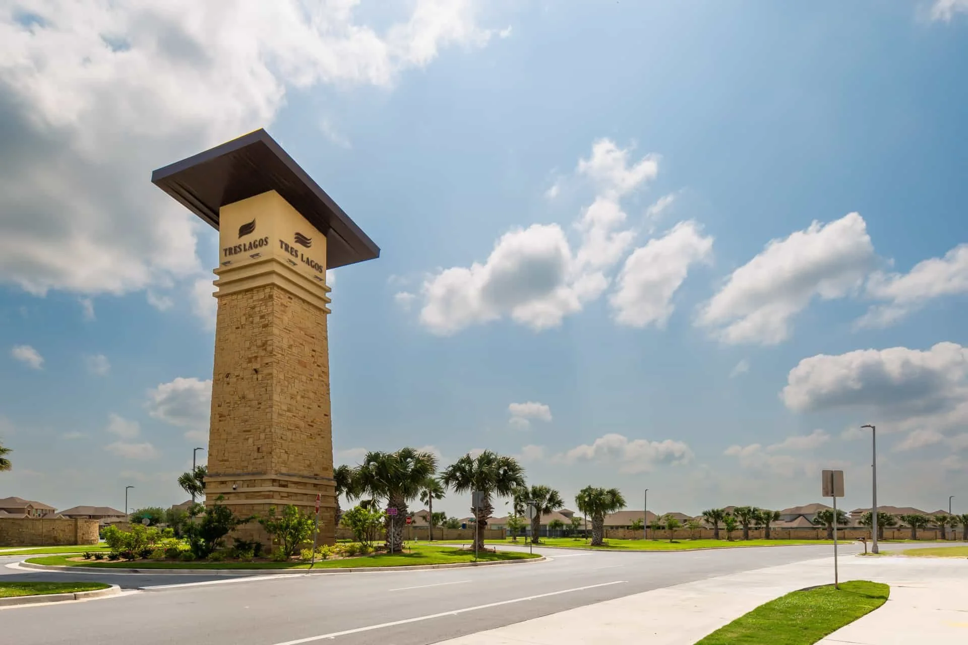 A tall stone entrance signpost with the word "Tres Lagos" and logo, under a black roof, set in a landscaped area with palm trees, a curved road, and a suburban neighborhood in the background under a partly cloudy sky.
