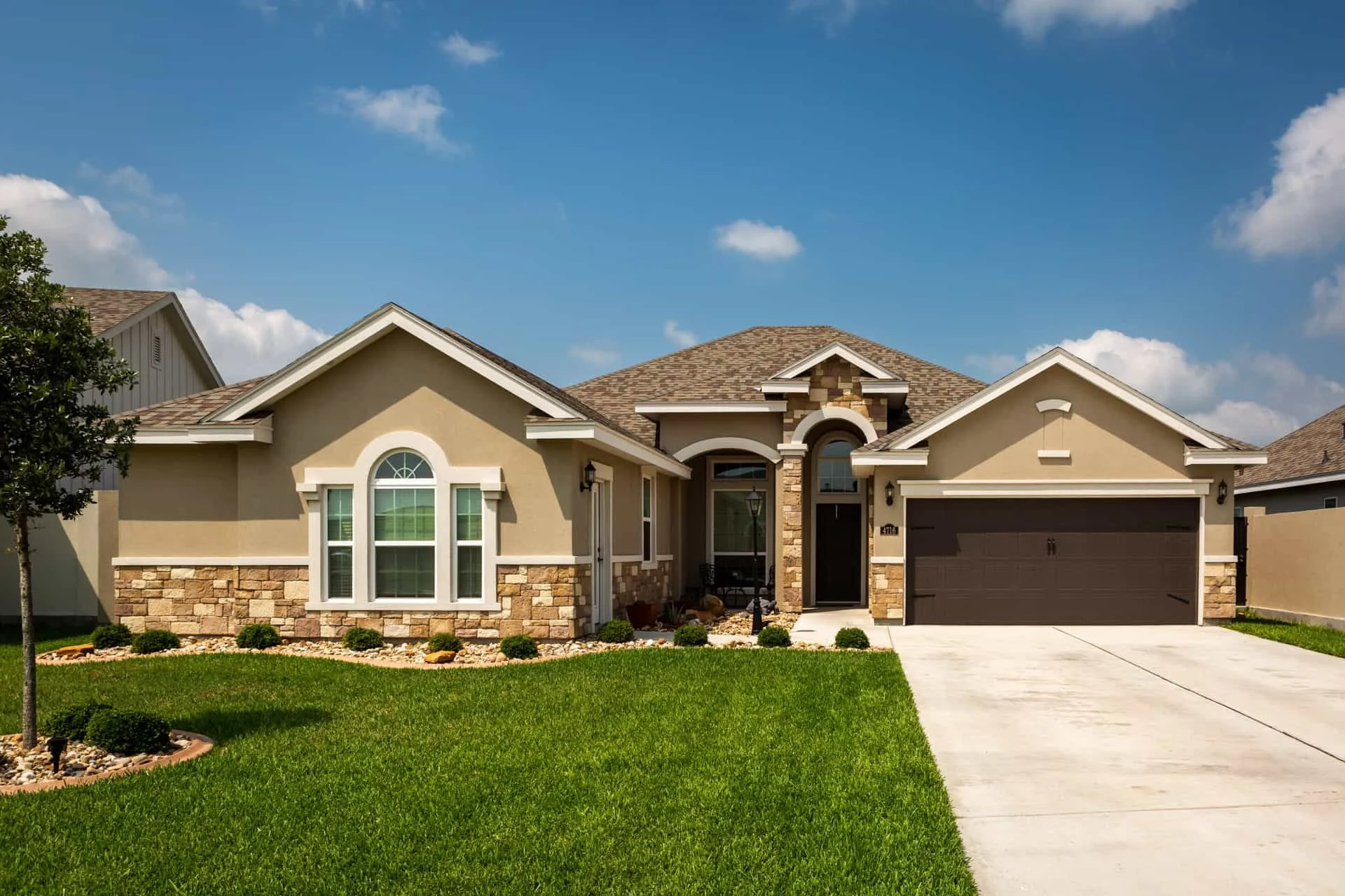 Front view of a single-family house with a well-maintained lawn, stone and stucco exterior, arched windows, and a two-car garage with a concrete driveway, under a partly cloudy blue sky.