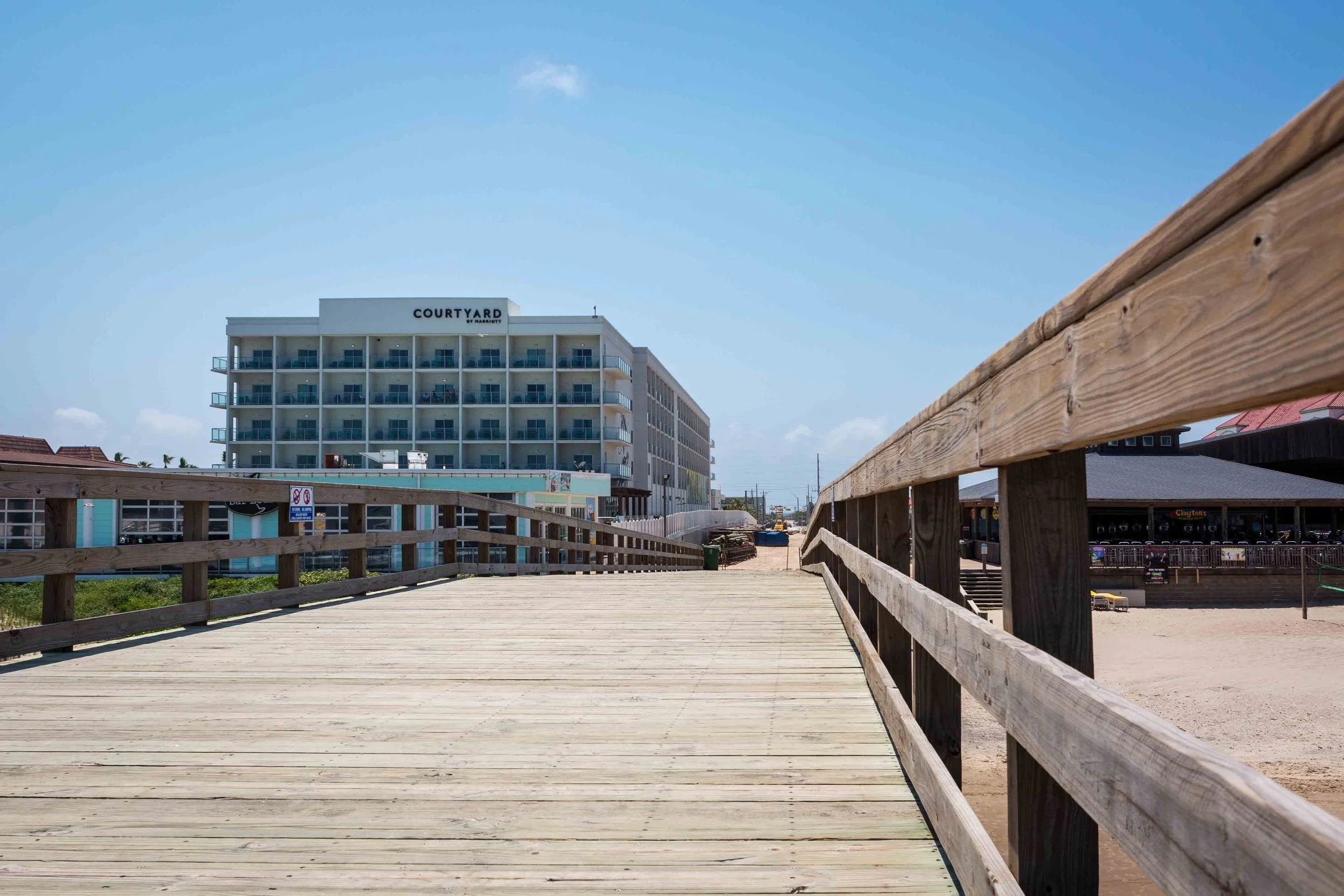 View of a wooden boardwalk on a beach leading toward a hotel building named 'Courtyard by Marriott' in the background with a blue sky and some clouds.