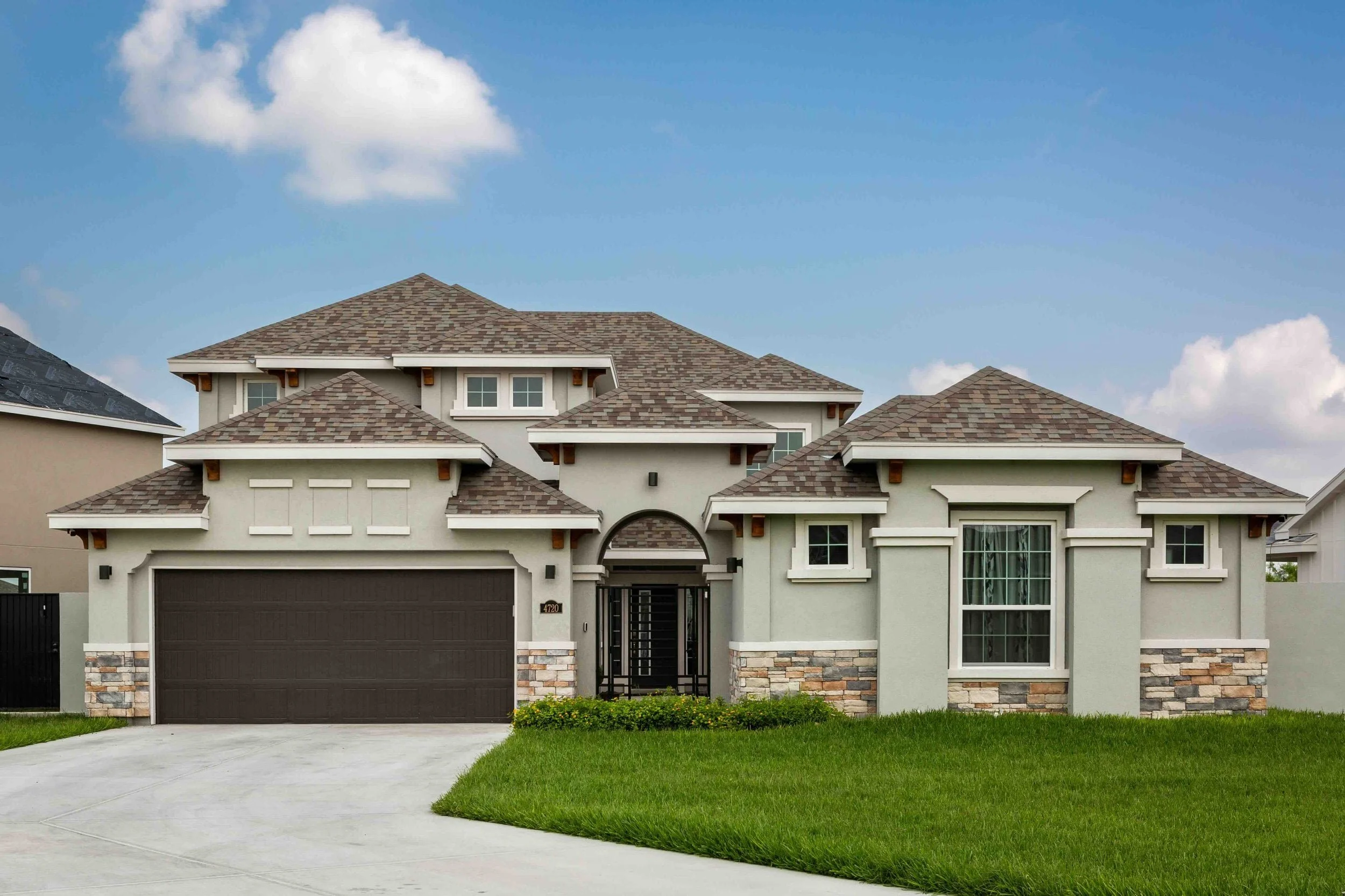 A modern two-story house with a brown shingle roof, a dark garage door, and a front yard with green grass and a concrete driveway. The house has multiple windows, some with white framing, and a stone veneer along the lower part of the exterior walls.