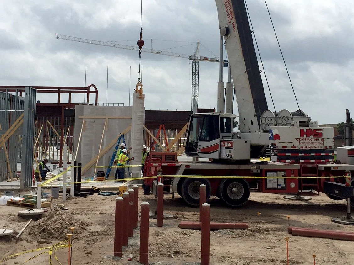 Construction site with workers and a crane lifting a large concrete wall segment. Construction materials and safety tape are visible, with a crane in the background.