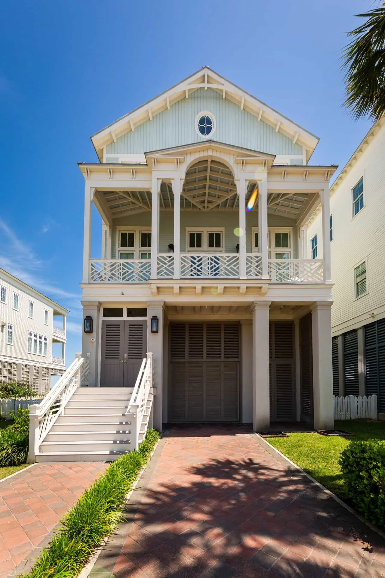 A tall, light-colored house with a gabled roof and a large balcony, located on a brick-paved path with green grass on each side, under a clear blue sky.