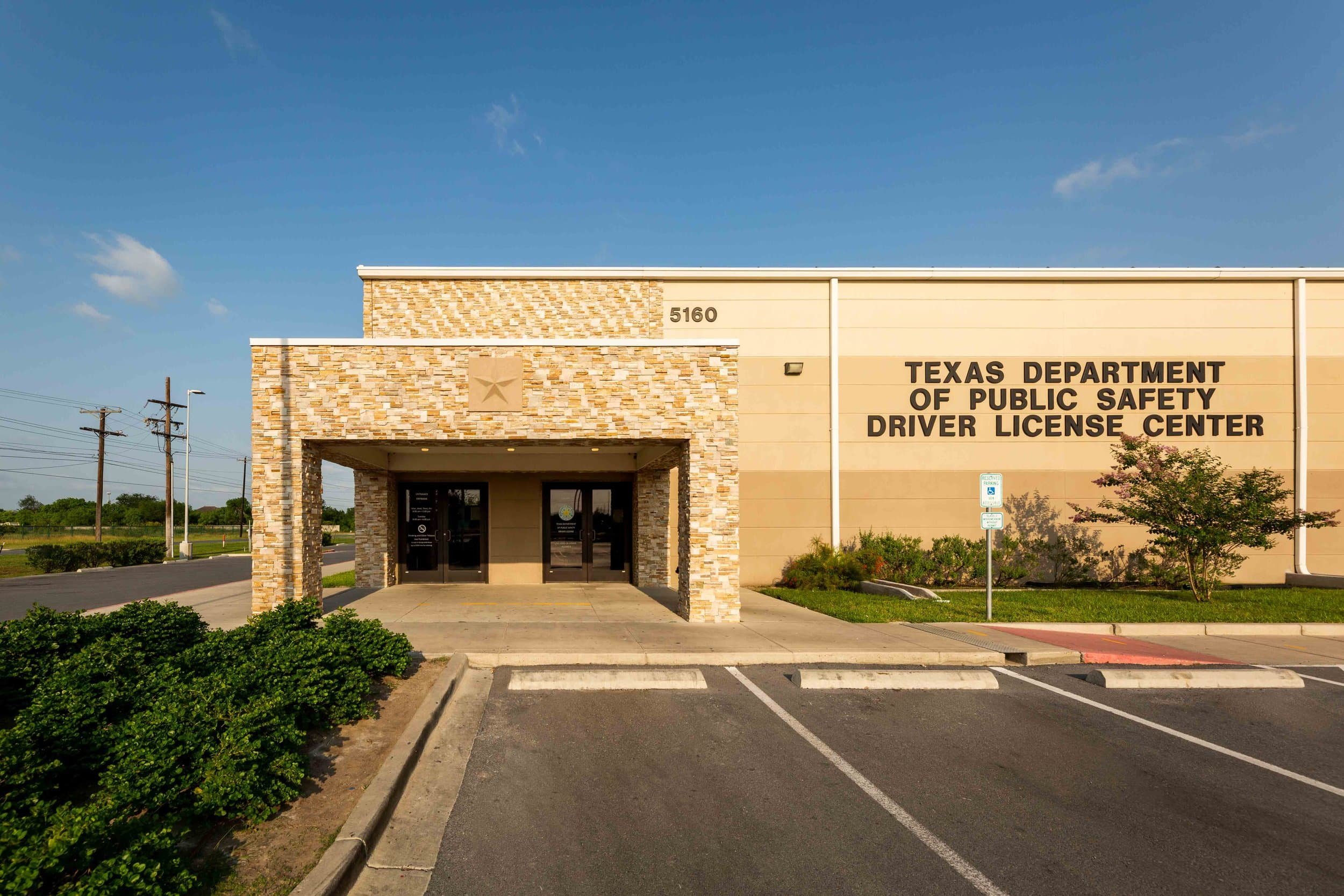 Front view of the Texas Department of Public Safety Driver License Center building with a stone and beige facade, parking spaces in the foreground, and power lines against a blue sky.