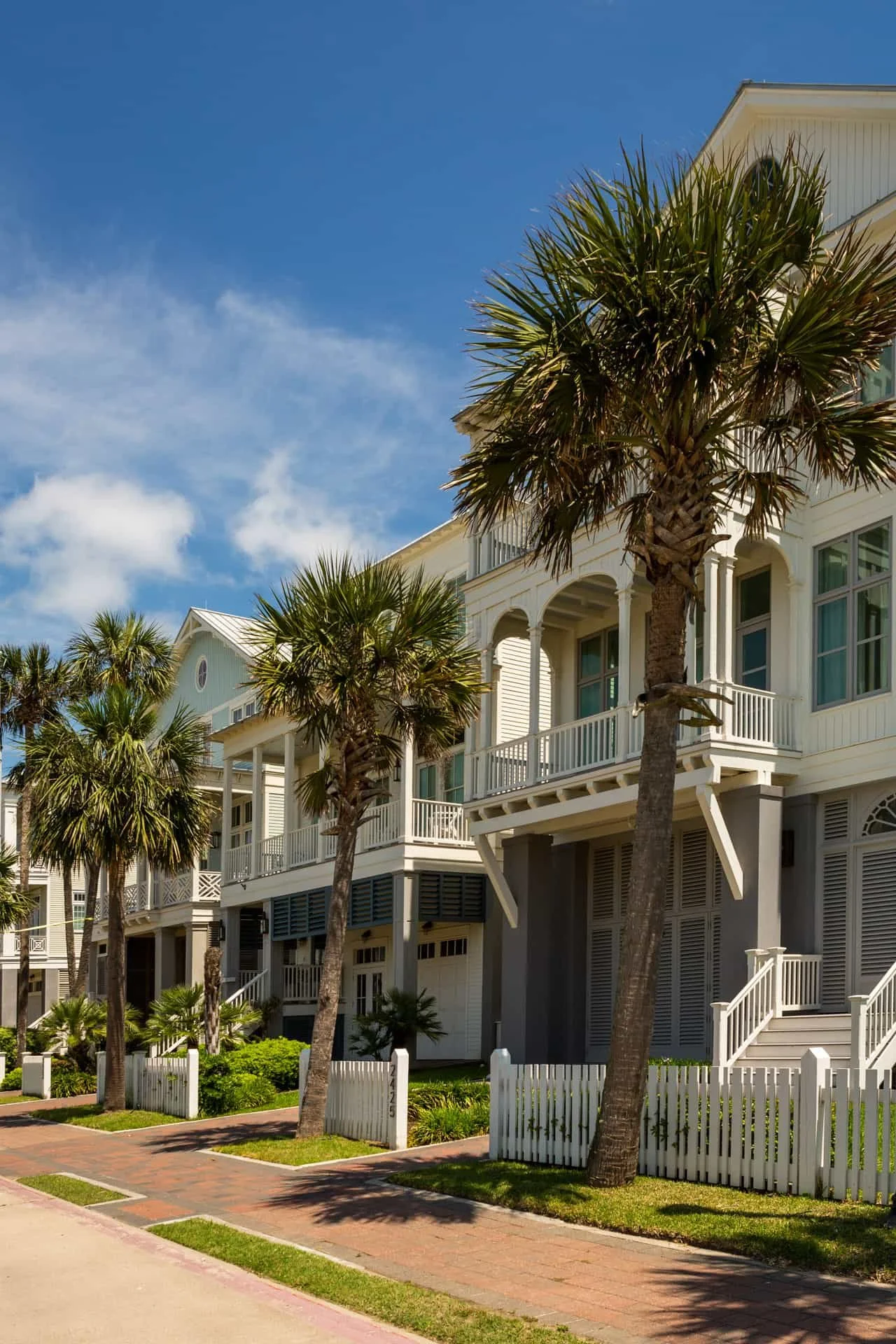 White beachfront houses with balconies and palm trees on a sunny day under a partly cloudy sky.