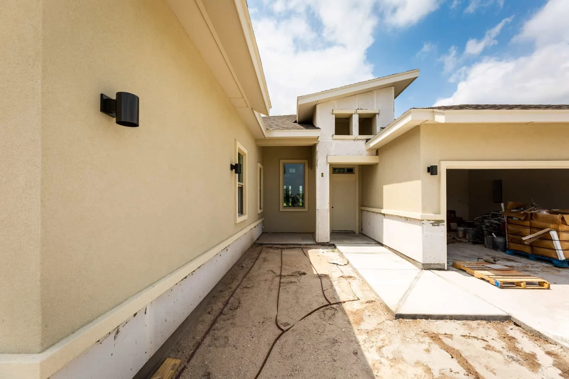 House exterior under construction, beige walls, black wall lights, and an open garage with construction materials inside