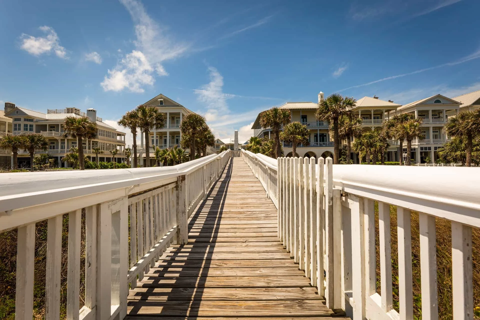 A wooden boardwalk leading to beachfront houses with palm trees under a partly cloudy blue sky.