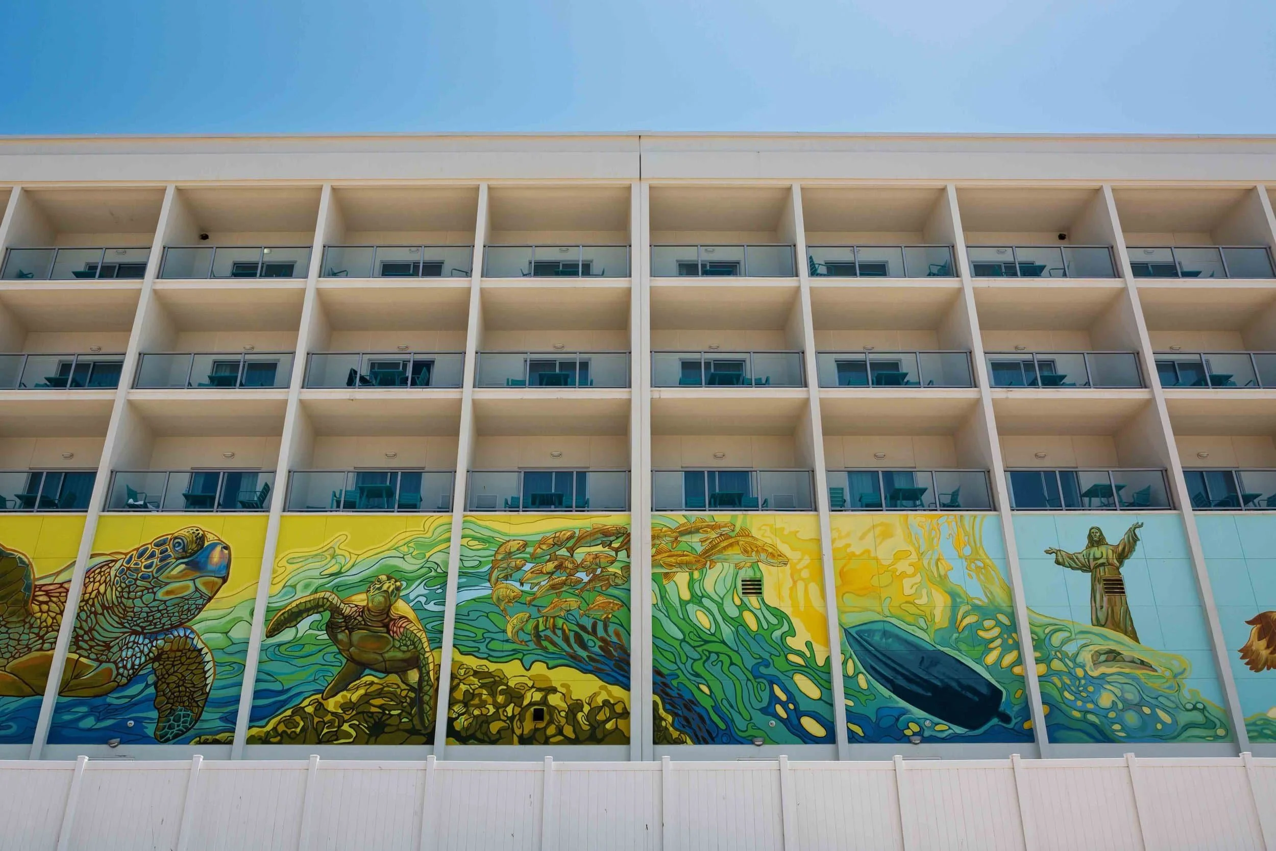Colorful mural of ocean animals, including turtles, fish, and a dolphin, painted on the side of a building beneath balconies with glass railings, against a clear blue sky.