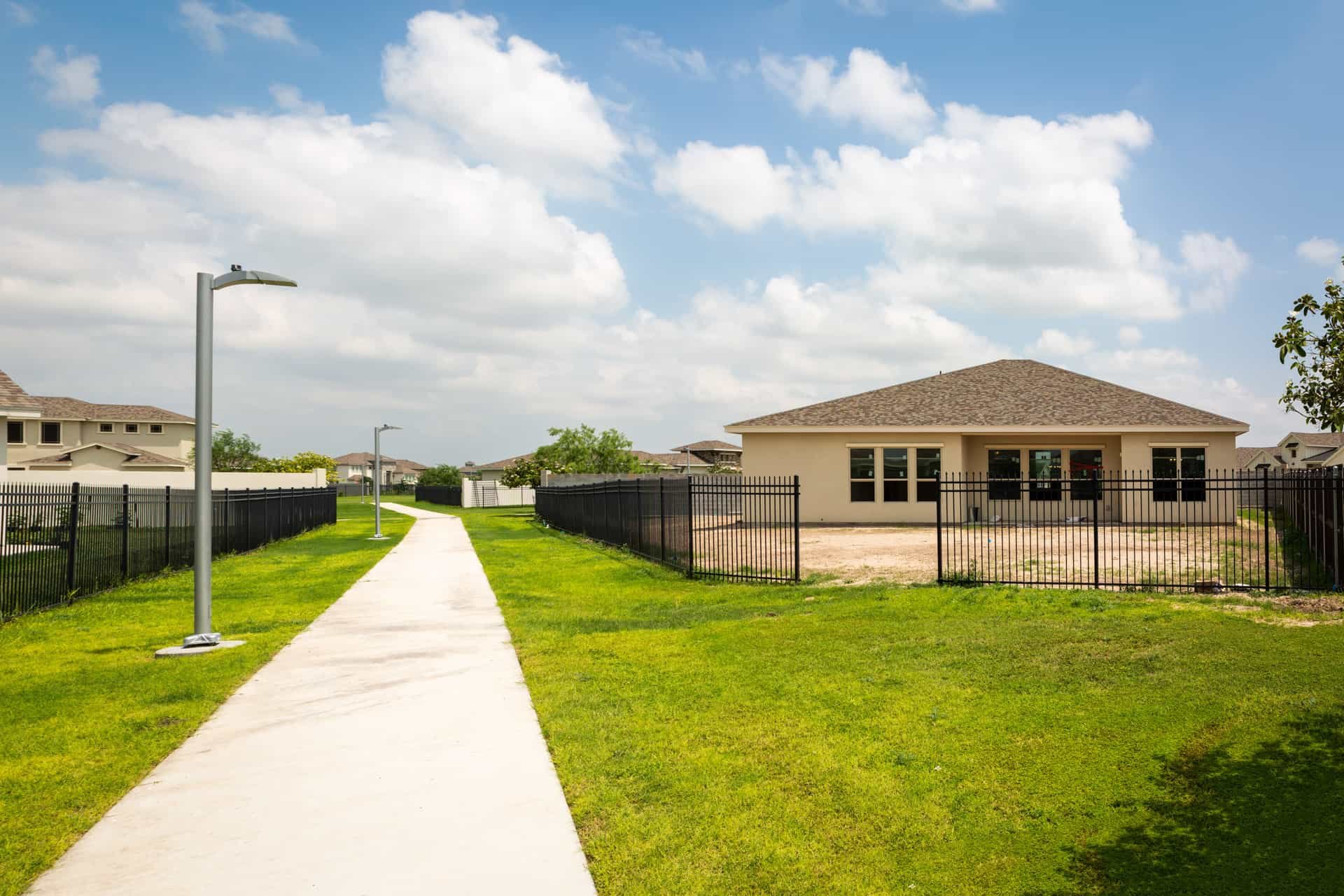A concrete sidewalk runs through a grassy area on a sunny day, with single-story houses in the background, a building under construction surrounded by a black fence, and tall street lamps along the sidewalk under a partly cloudy sky.
