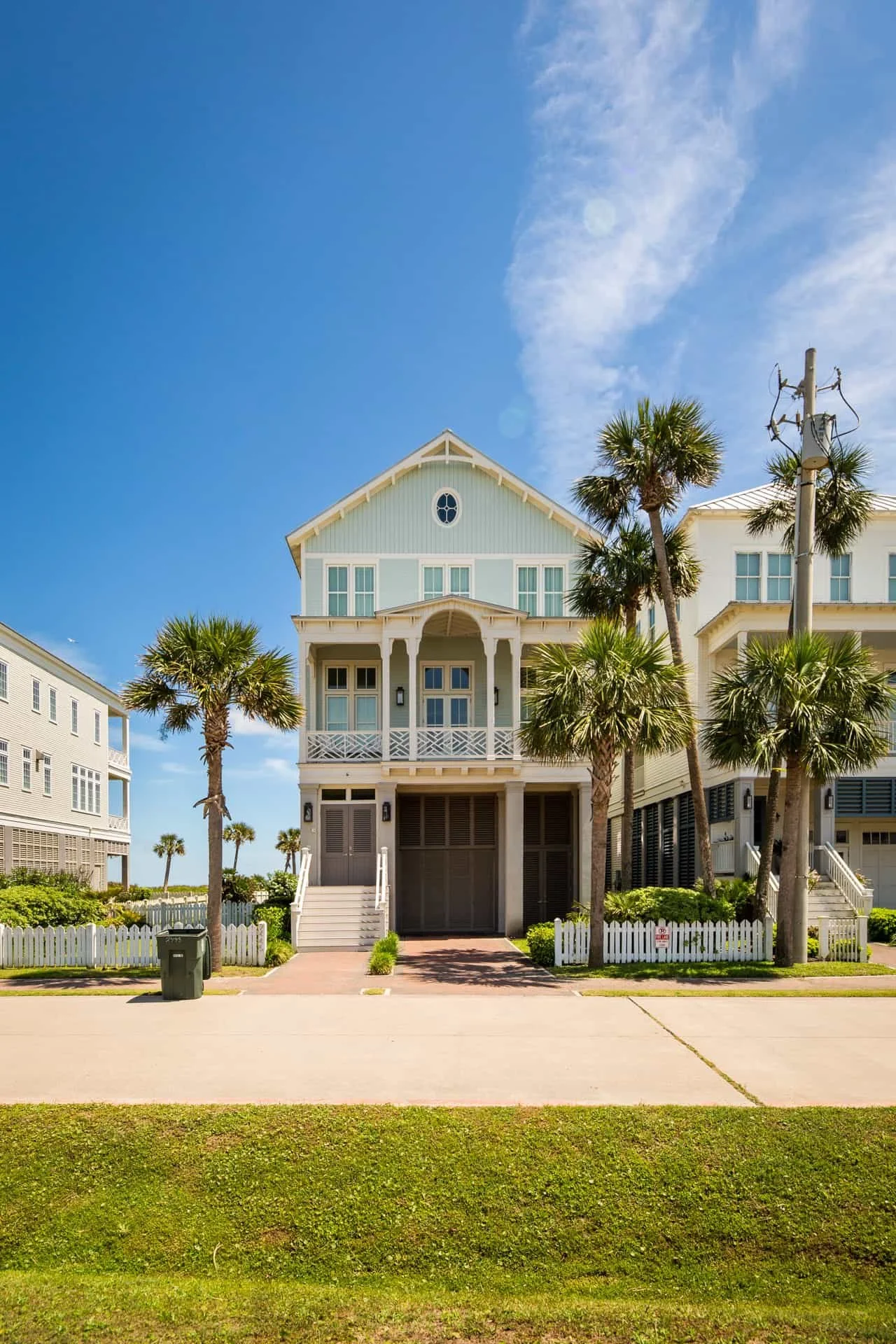 A large beach house with a pastel blue exterior and white trim, surrounded by palm trees, under a bright blue sky with scattered clouds.