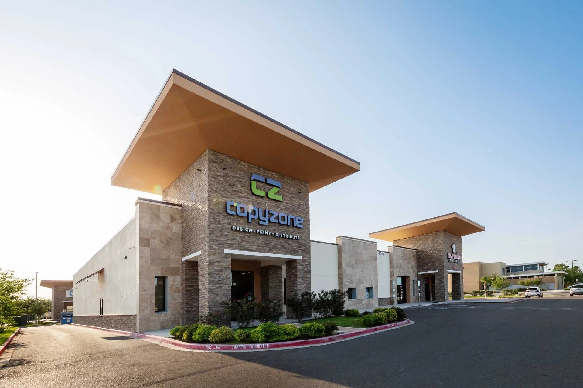 Exterior of C2 Copyzone retail store in a shopping plaza with a large, modern canopy roof, brick facade, and sign displaying services like design, print, and distribute. Clear sky and parking lot in the foreground.