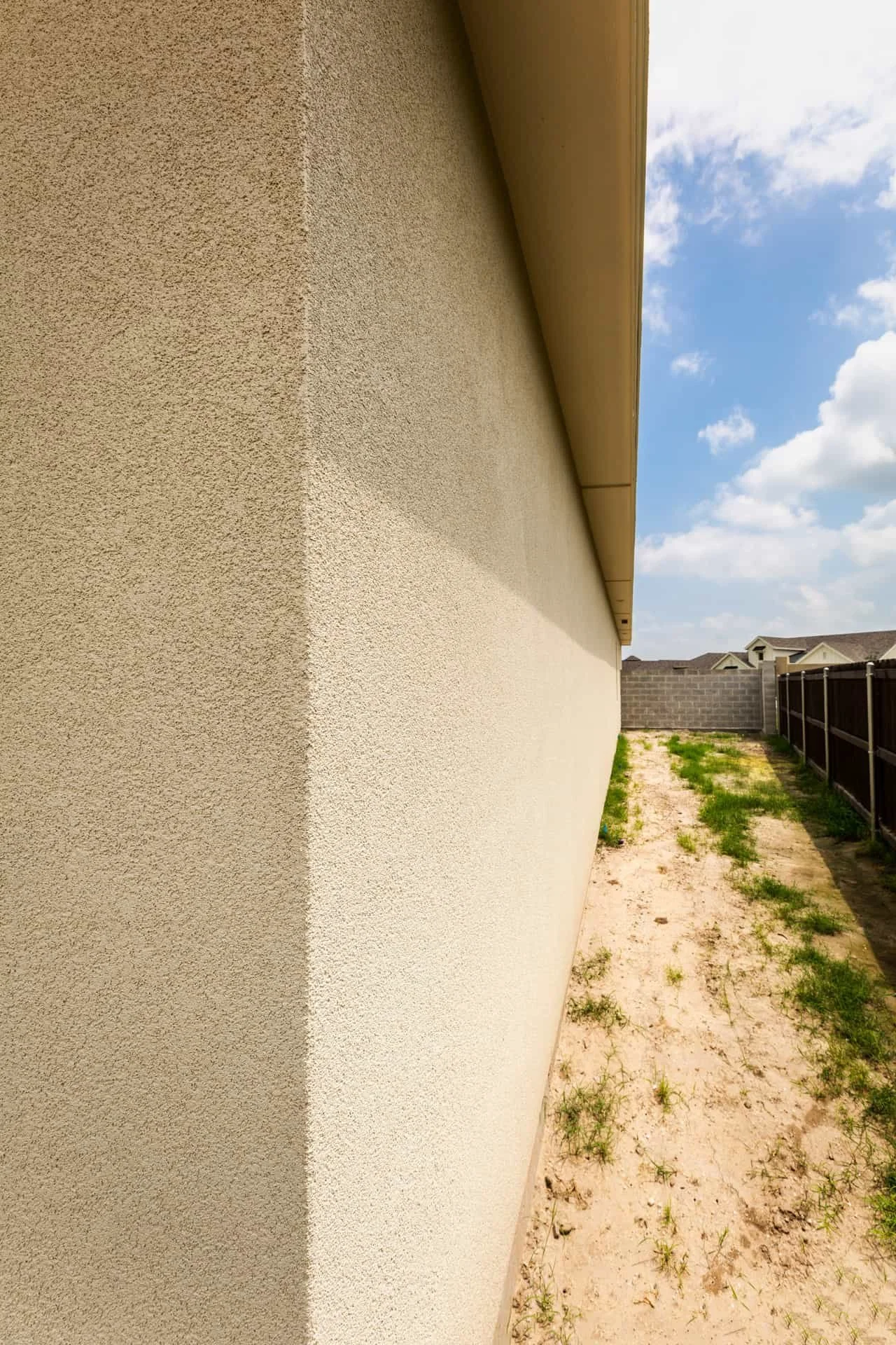 Side yard view of a house with textured beige exterior wall, dry grass, and a black fence, under a blue sky with some clouds.