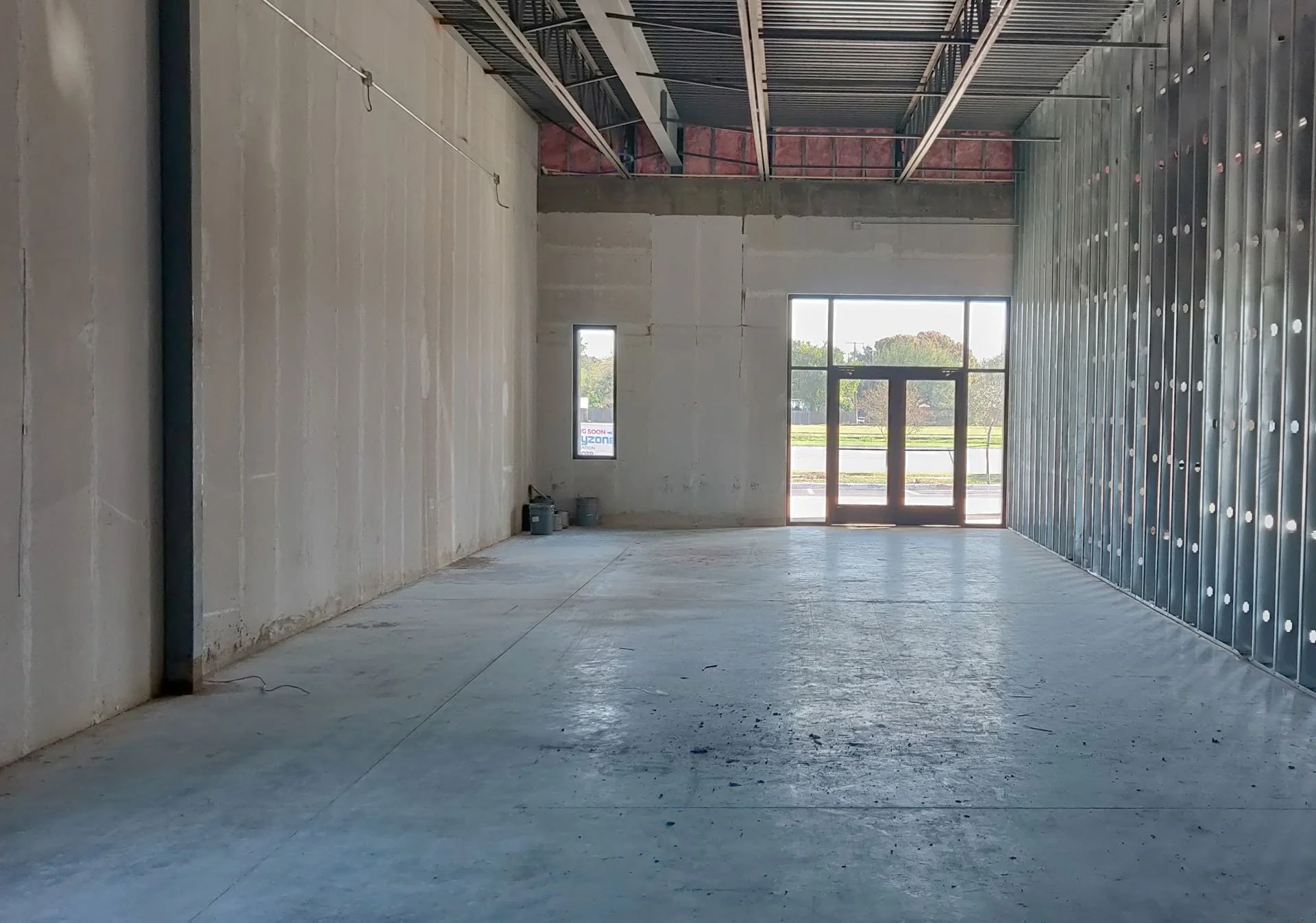 Interior of a building under construction with unfinished walls, large front window, and a side window, concrete floor, and exposed metal framing on the ceiling.
