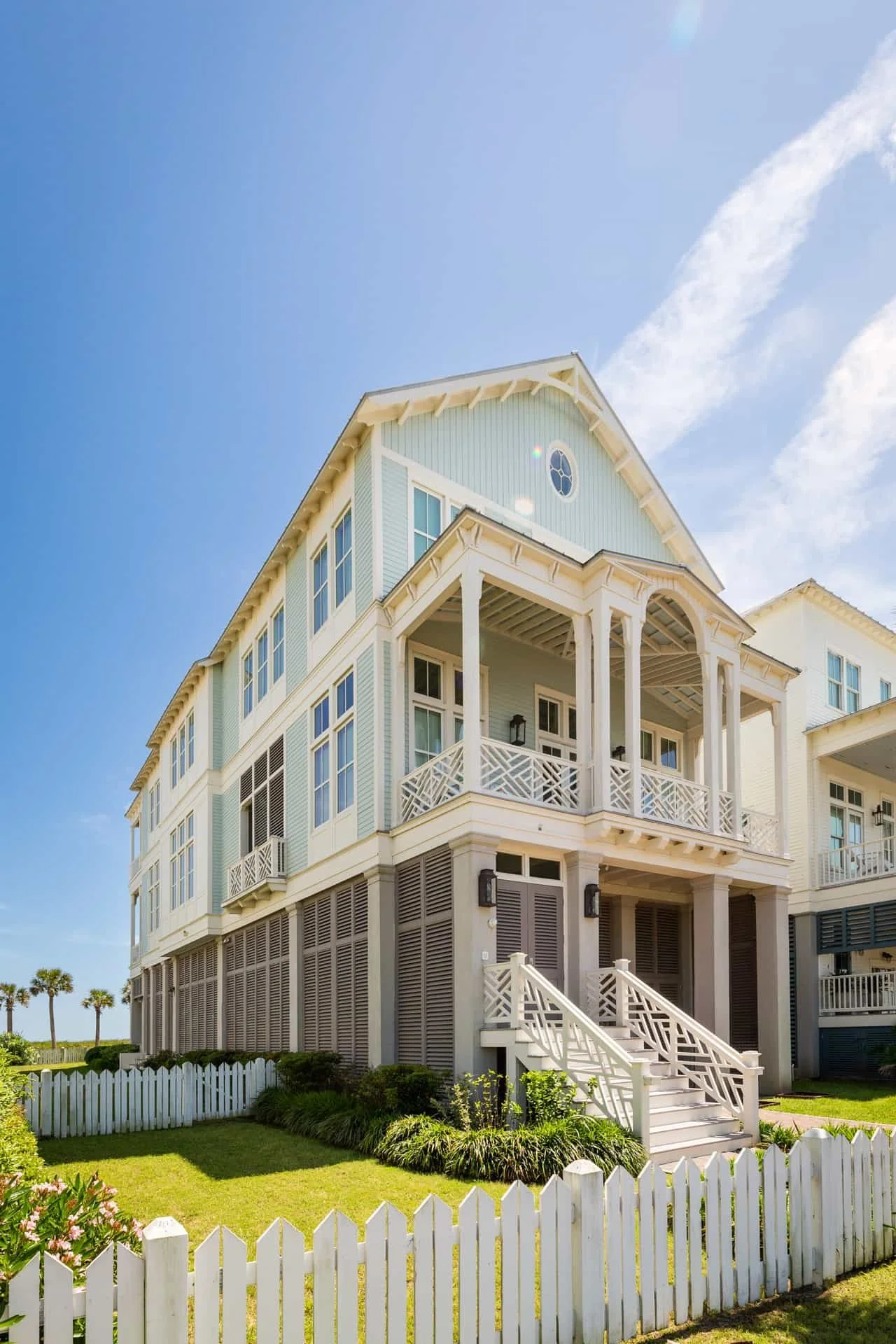A white multi-story house with a wrap-around porch, set against a bright blue sky. The house is surrounded by a white picket fence and green grass.
