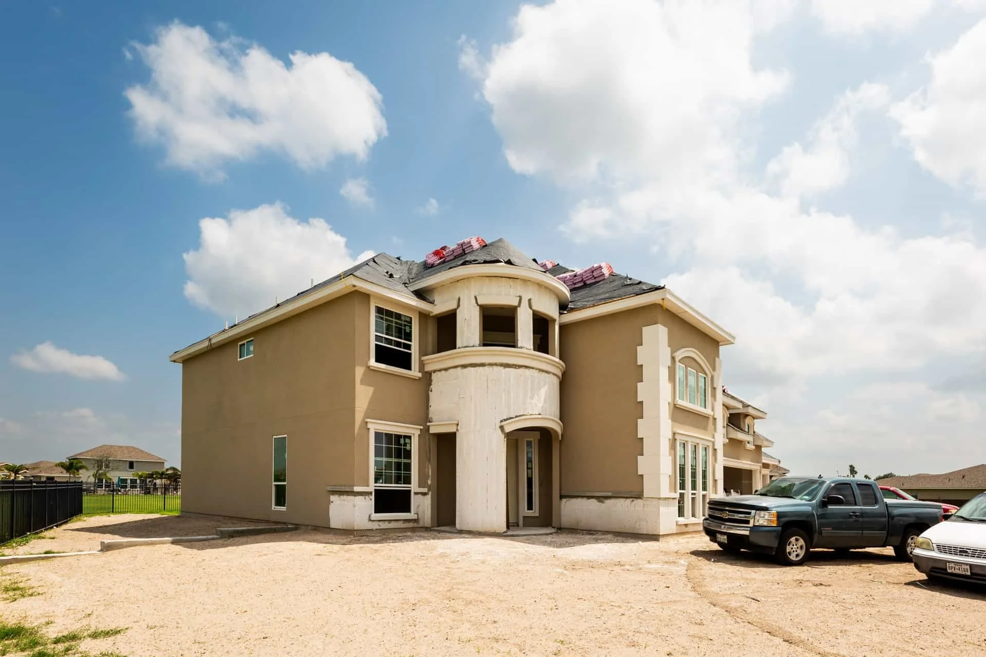 A two-story beige house under construction with a curved balcony and several windows, parked cars in front, and a partly cloudy sky.