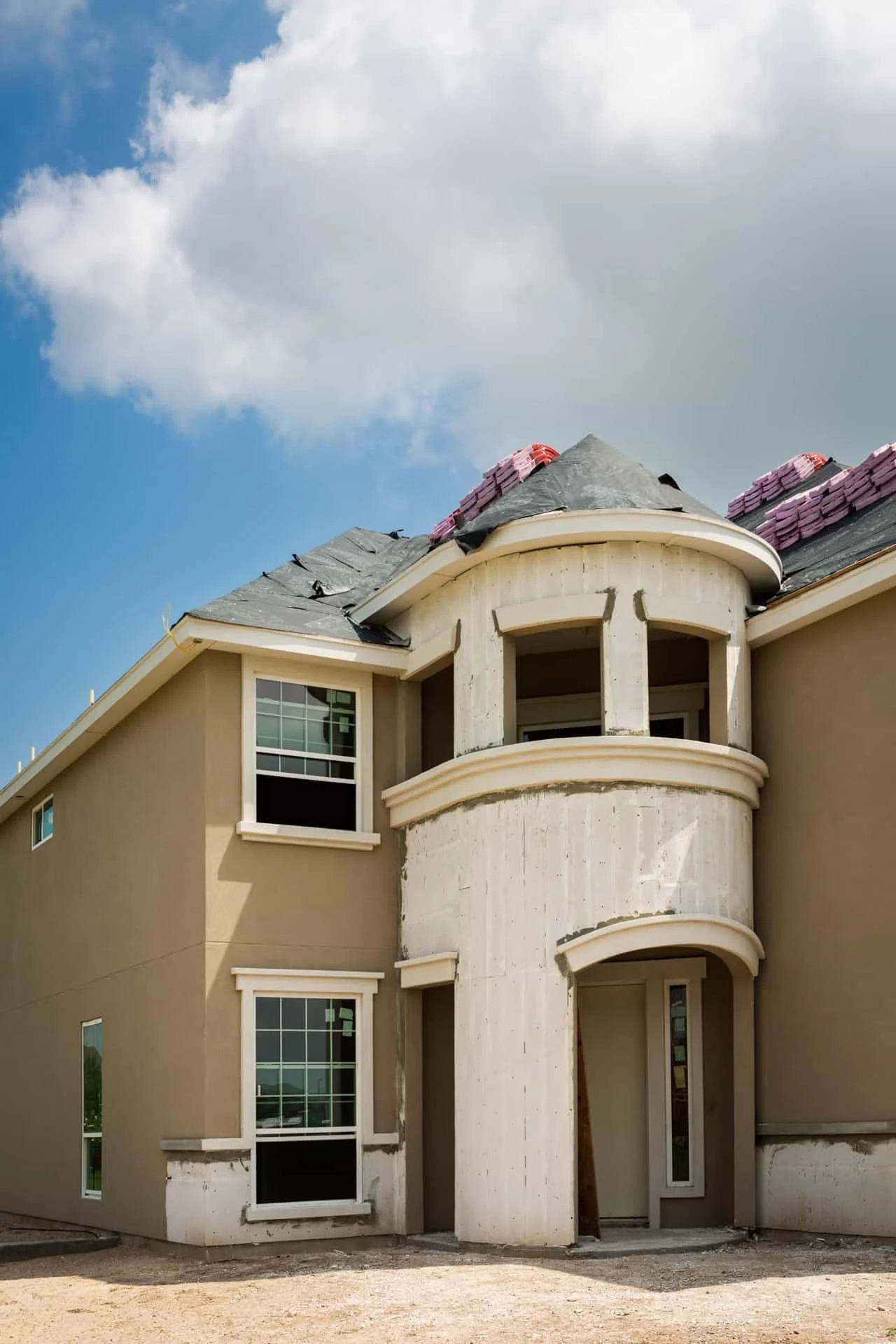 A house under construction with an unfinished curved tower, beige walls, and a partially completed roof, with a cloudy sky above.