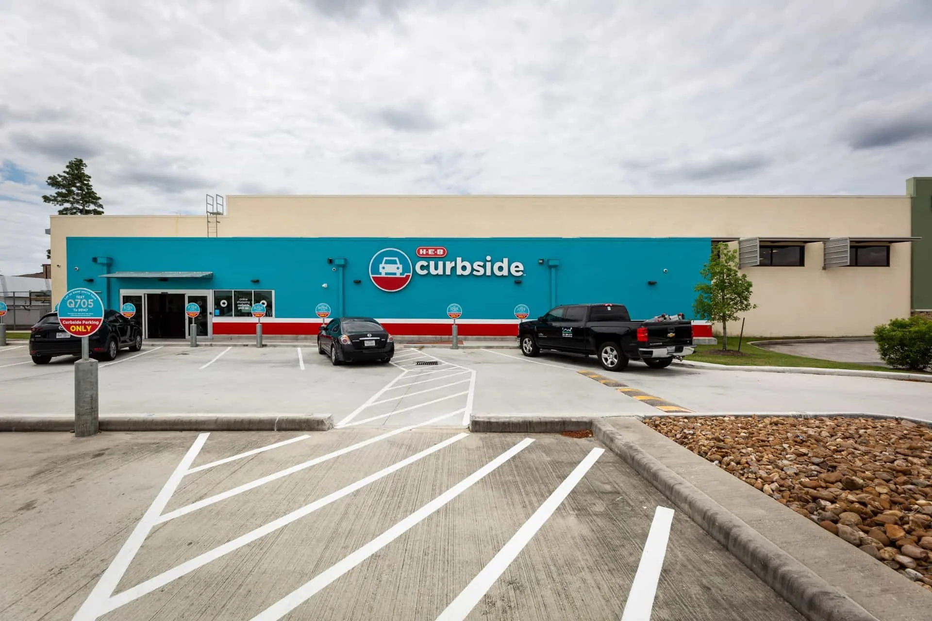 Autoclaved Aerated Concrete (AAC) I Exterior view of a H-E-B Curbside grocery pickup location with parking spaces, some cars parked, a building with a blue and beige facade, and a cloudy sky.