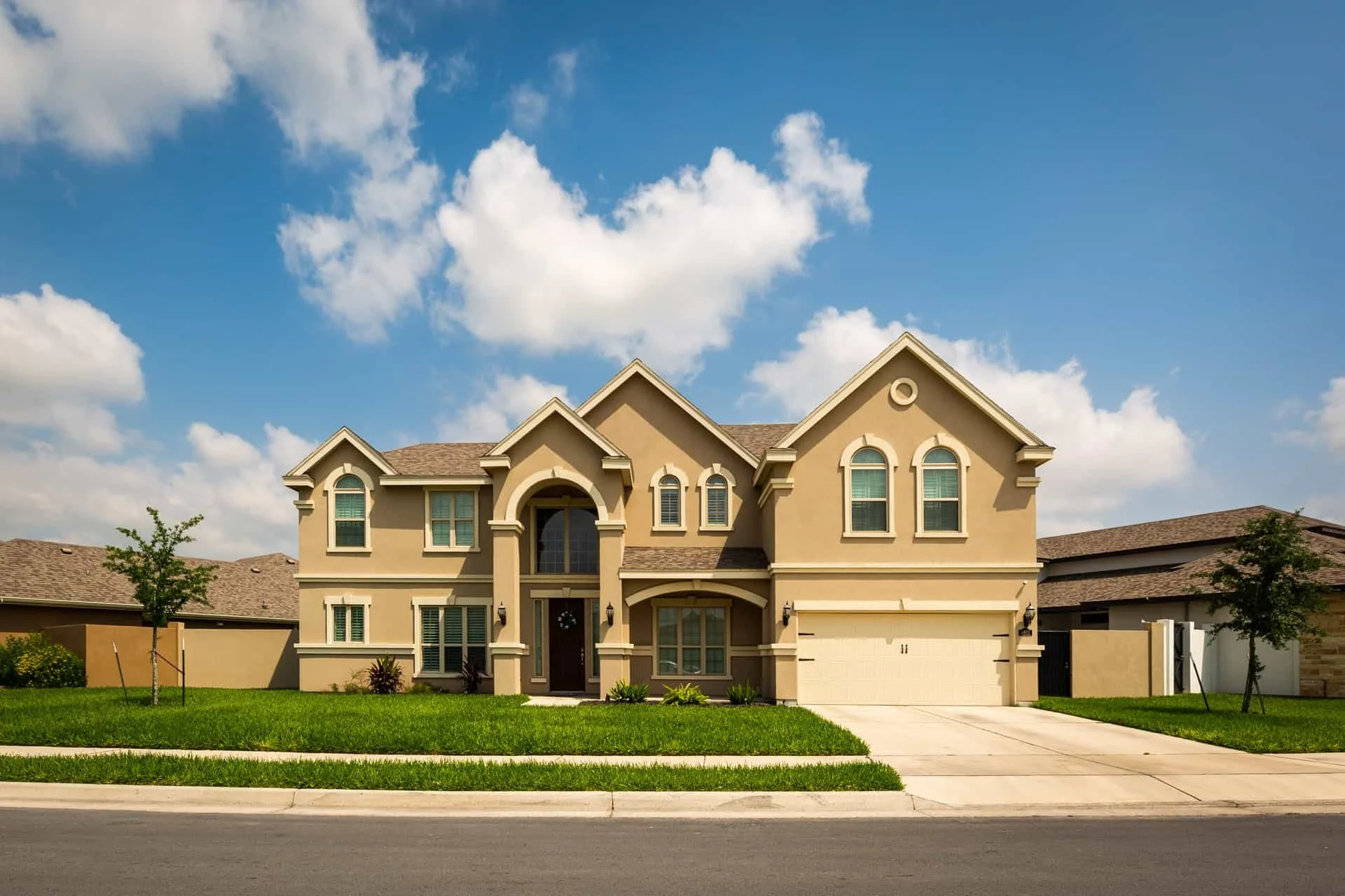 A two-story beige house with a garage, front lawn, and clear blue sky with clouds.