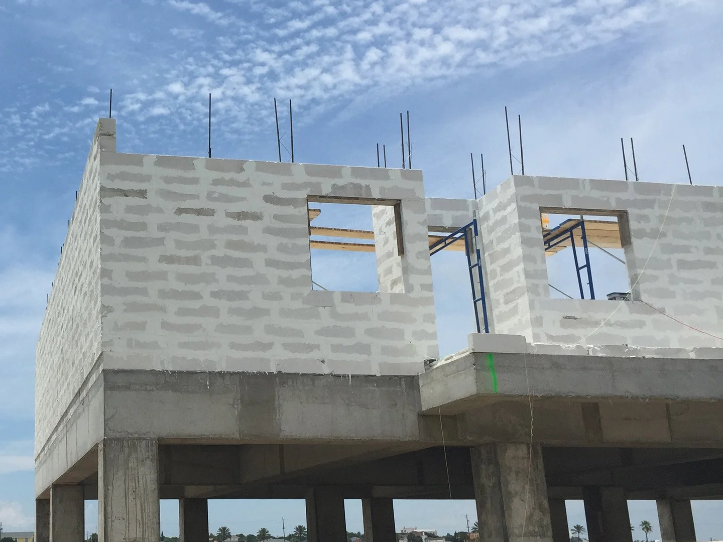 Construction site of a building with white brick walls, window openings, and concrete supports, under a partly cloudy sky.
