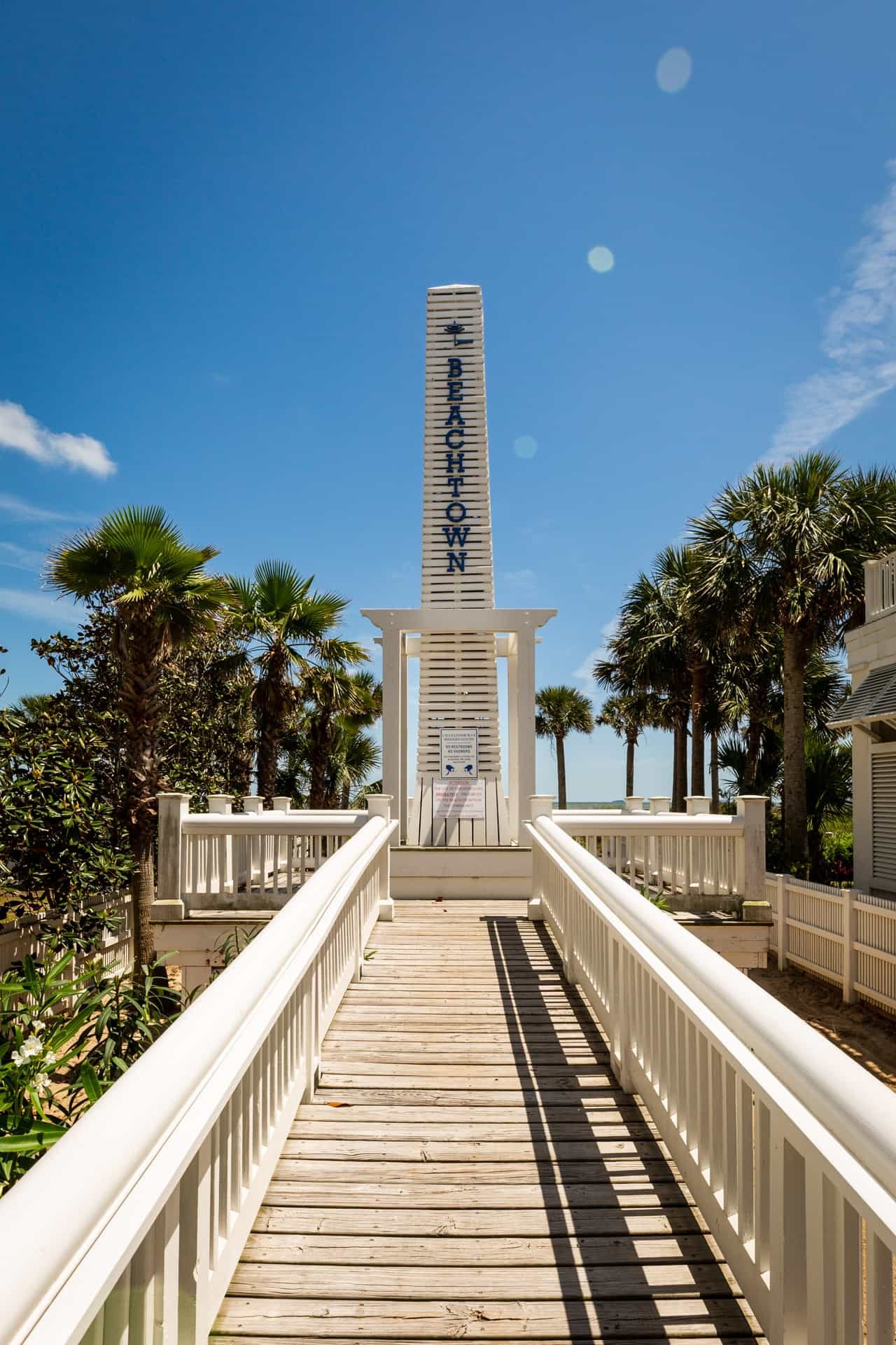 A white wooden walkway leading to a white structure with a tall sign that reads "BEACH TOWN" against a blue sky with some clouds, surrounded by palm trees.