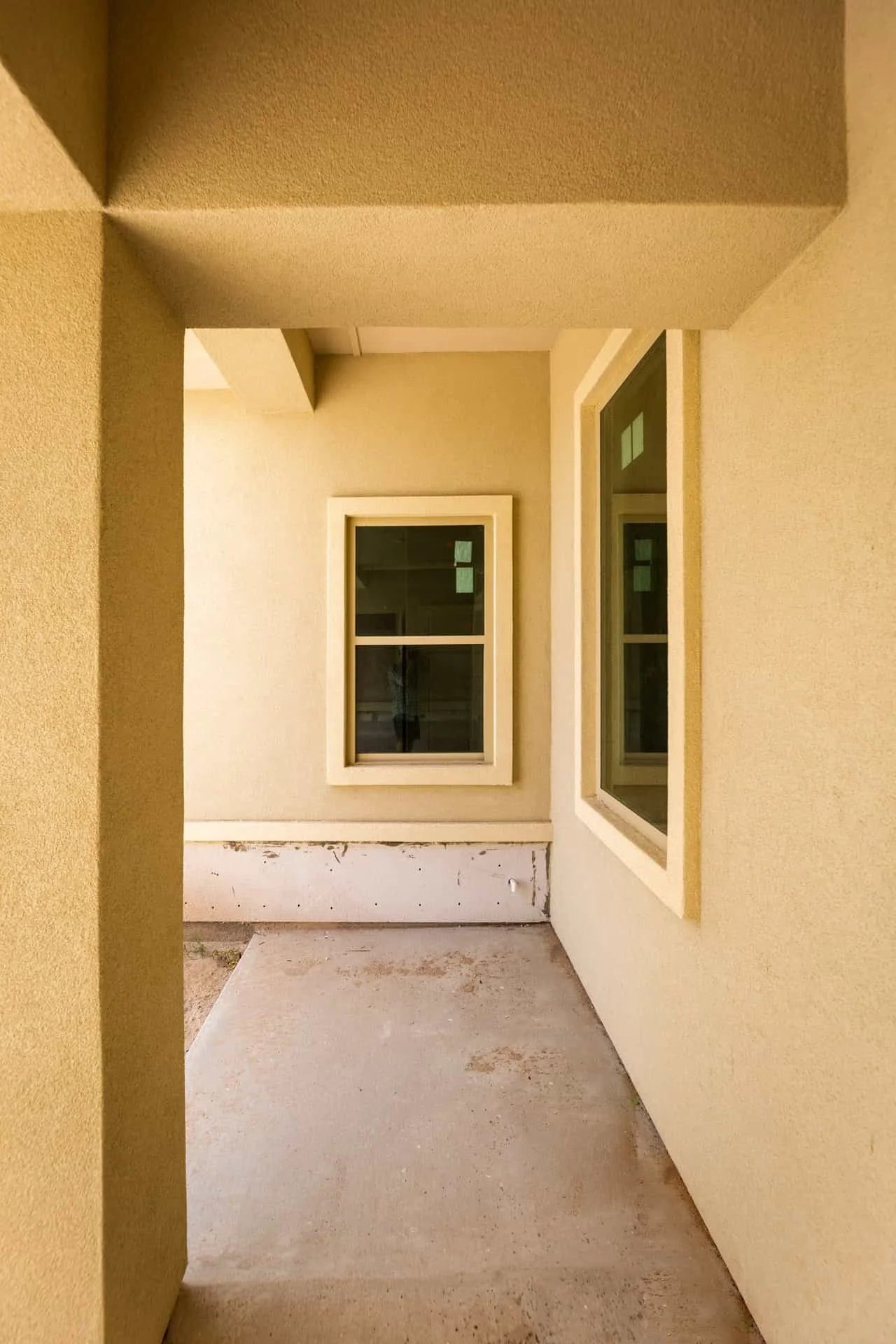 Exterior view of a partially finished patio with two white-framed windows on a yellow stucco wall and a concrete floor.