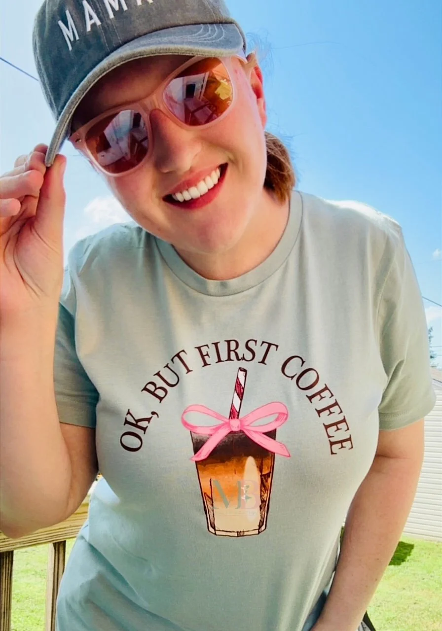 A smiling woman wearing a blue cap and pink sunglasses, adjusting her cap, standing outdoors on a sunny day. She is wearing a beige T-shirt with a graphic of a coffee cup adorned with a pink bow and the phrase 'OK, BUT FIRST COFFEE'.