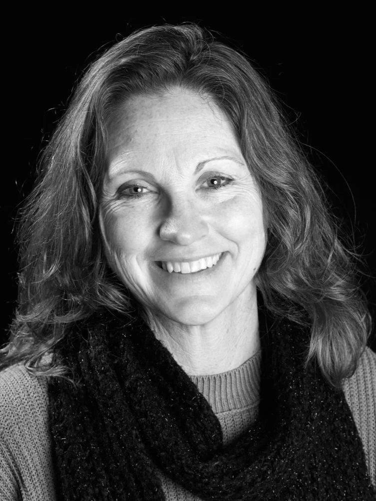 Black and white portrait of a middle-aged woman with wavy hair smiling, wearing a sweater and a scarf, against a dark background.