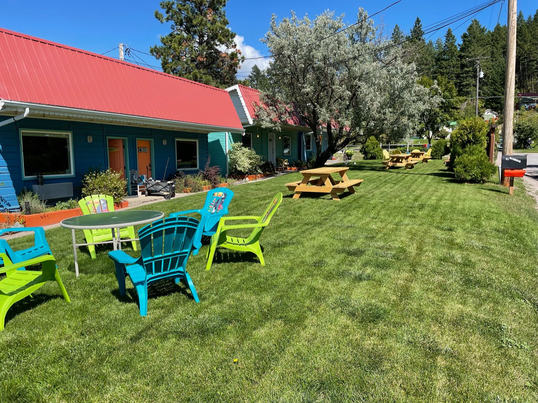 Outdoor seating area with colorful chairs and picnic tables on a green lawn next to a blue building with a red roof.