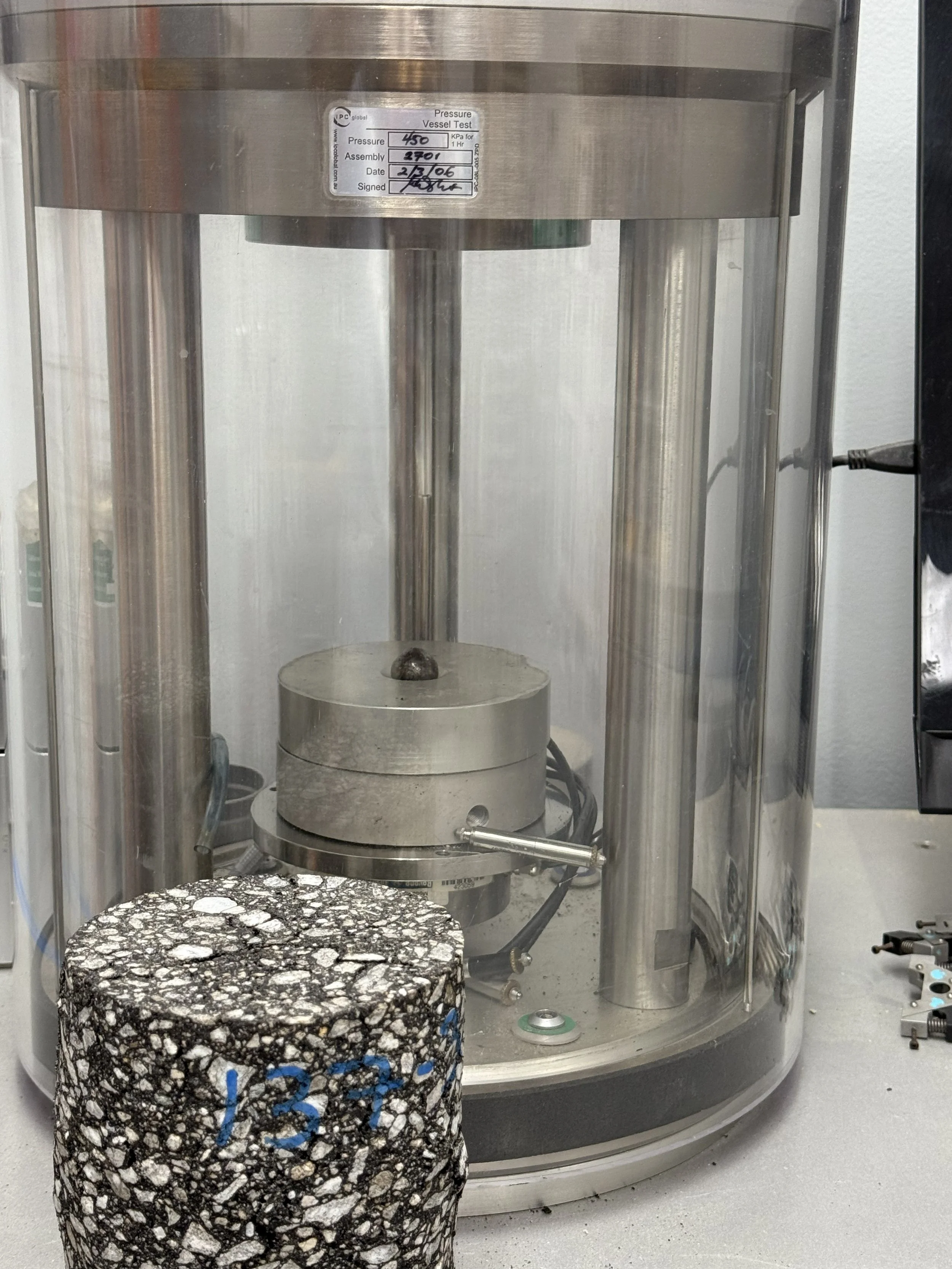 An AMPT testing device for modulus in a glass case, with a pressure test label on top, and a large cylindrical rock sample with numbers written in blue in front of it.
