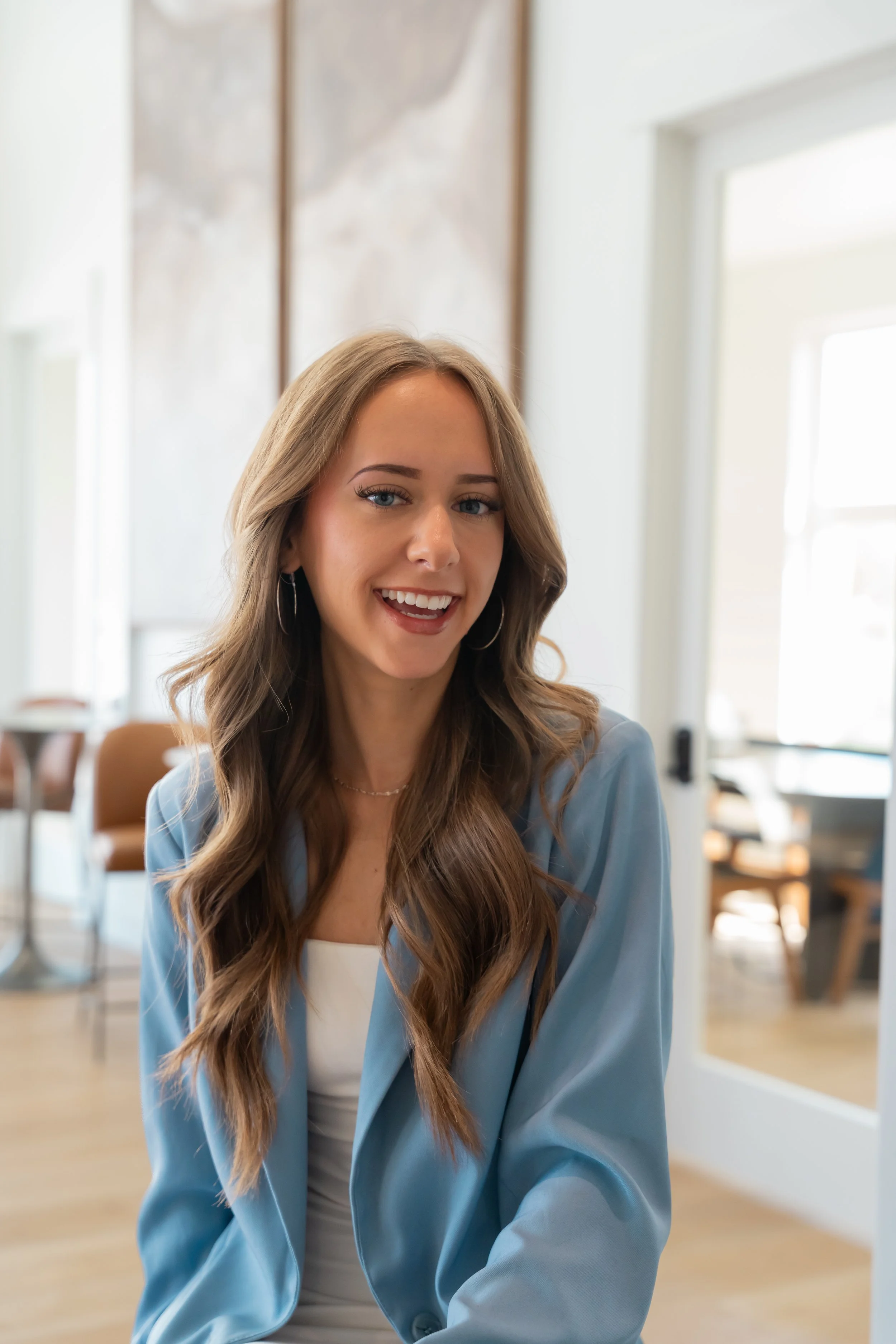 A young woman, Brianna Daniels with long wavy hair, blue eyes, and hoop earrings smiling in a bright indoor setting.