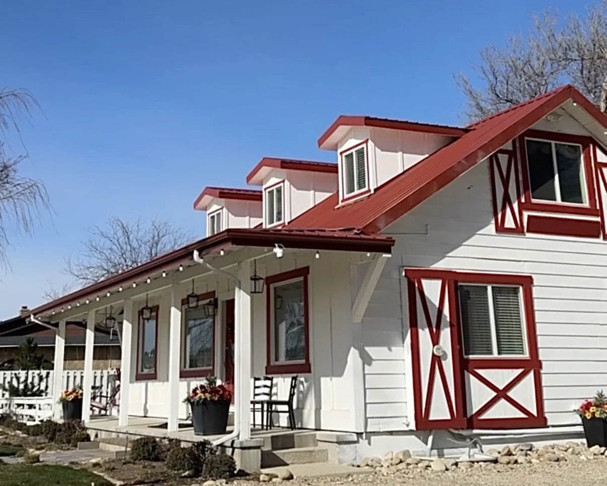 White house with red accents, red roof, and dormer windows, front porch and flower pots, under a clear blue sky.